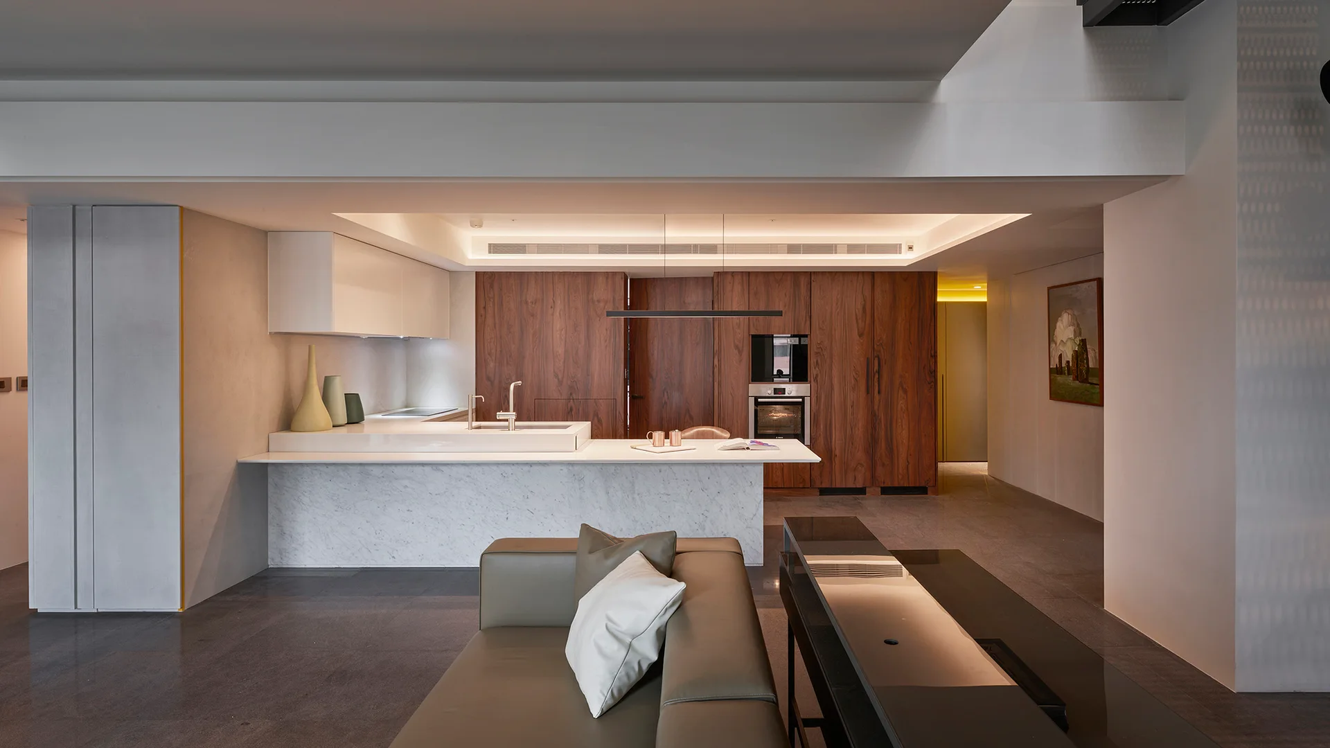 An open-concept living space features a white marble kitchen island, sleek walnut cabinets in the background, and a modern leather sofa in the foreground.