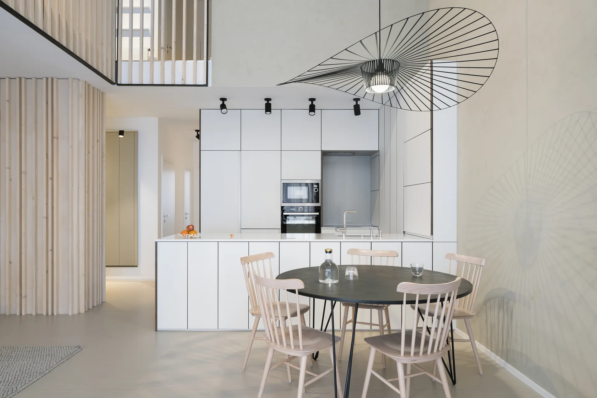 A modern Scandinavian kitchen features matte white cabinets, a light wood slat wall, and a round black dining table under a large wire pendant light.