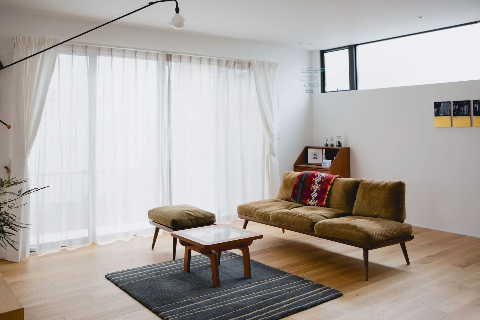 A minimalist living room features an olive green corduroy sofa, a matching ottoman, a low wooden coffee table, and sheer white curtains.