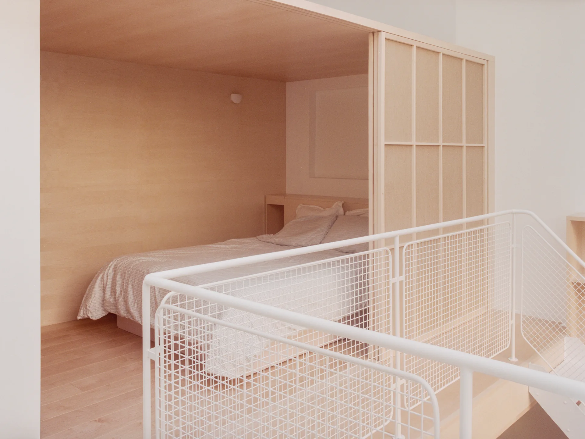 A light wood built-in sleeping nook containing a bed sits behind a white wire grid railing.