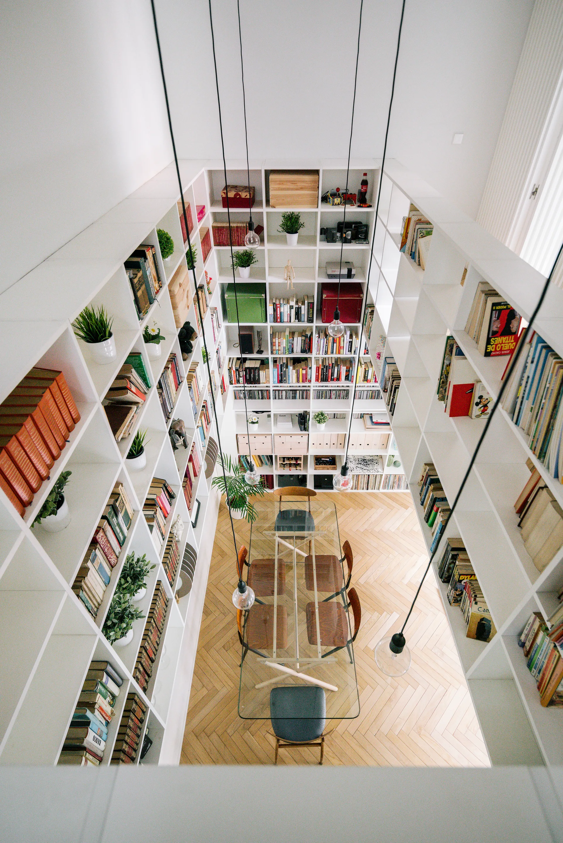 A high-angle shot of a dining area with a glass table, enclosed by towering white built-in bookshelves on herringbone wood floors.