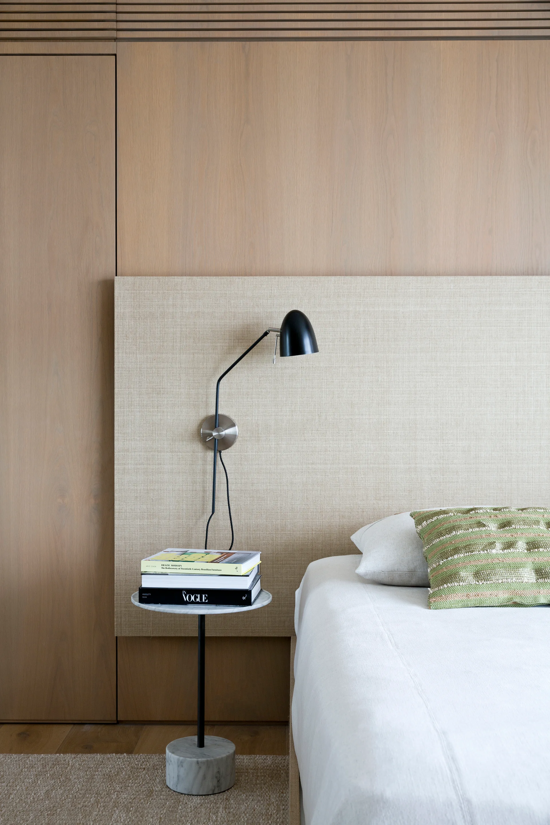 A modern minimalist bedroom featuring light wood wall paneling, an upholstered beige headboard, a black reading lamp, and a small marble side table.