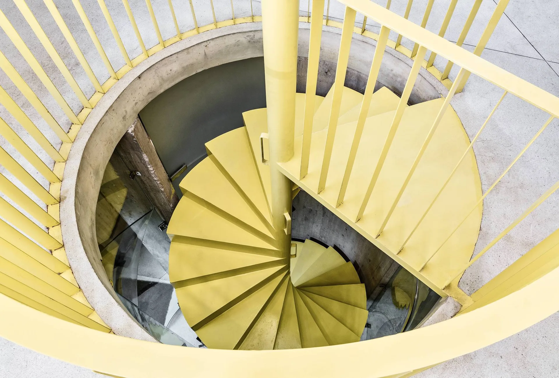 A high-angle overhead view of a bright yellow metal spiral staircase descending inside a circular concrete wall enclosure.