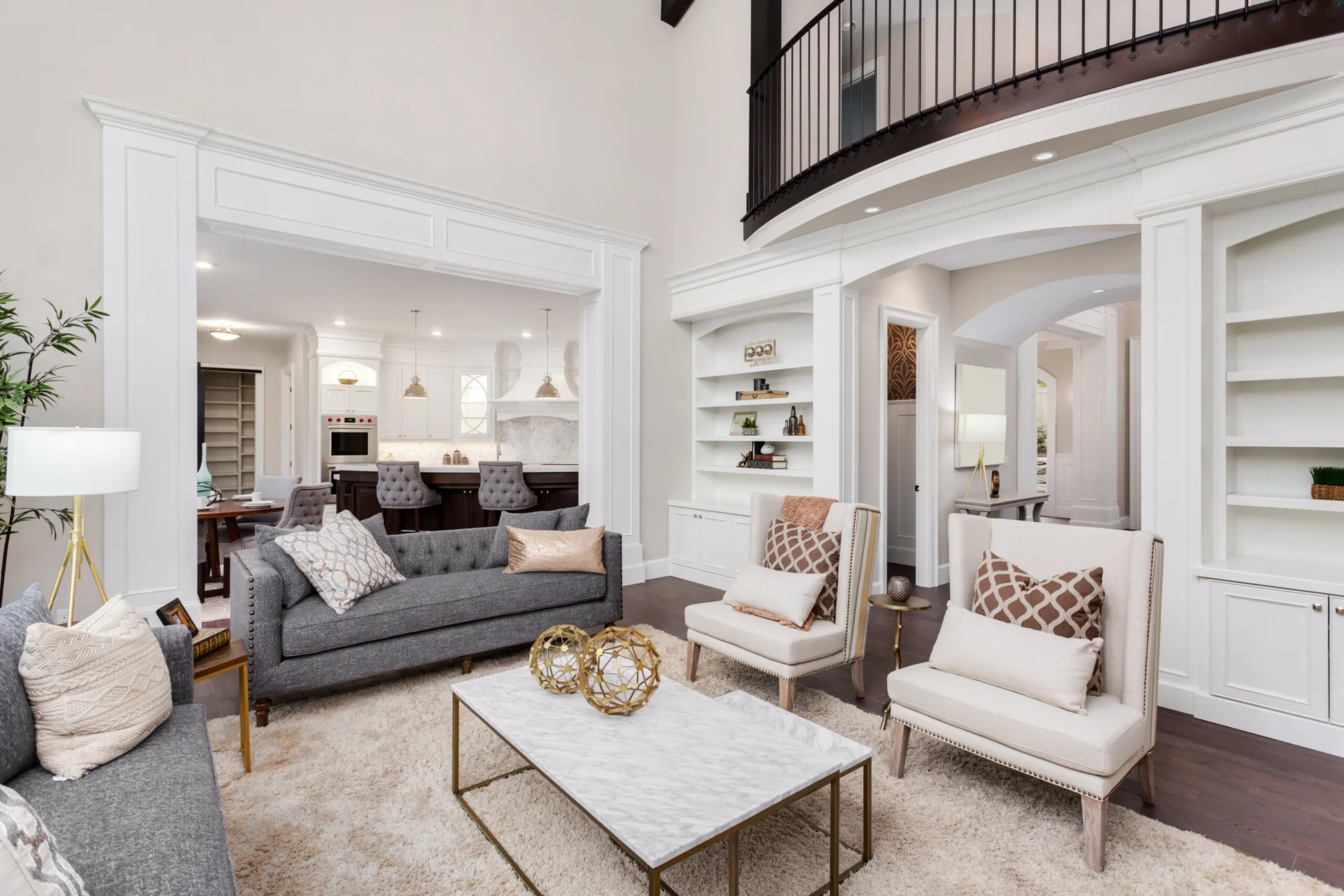 A transitional living room features a grey sofa, beige armchairs, a marble coffee table, and white built-ins opening to a kitchen.