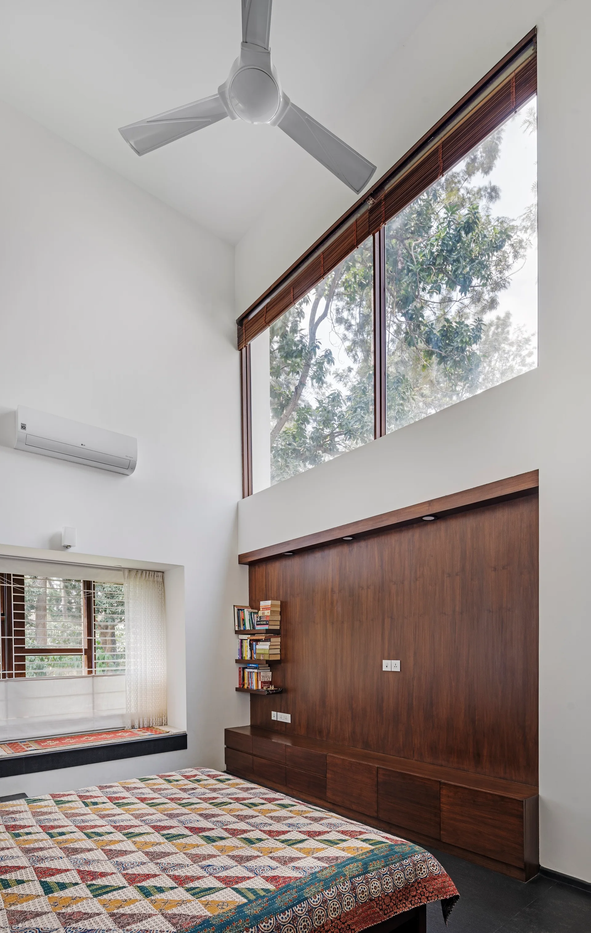 A modern bedroom features a high ceiling, a large upper window, a rich wood accent wall with low cabinets, and a bed with a colorful geometric quilt.