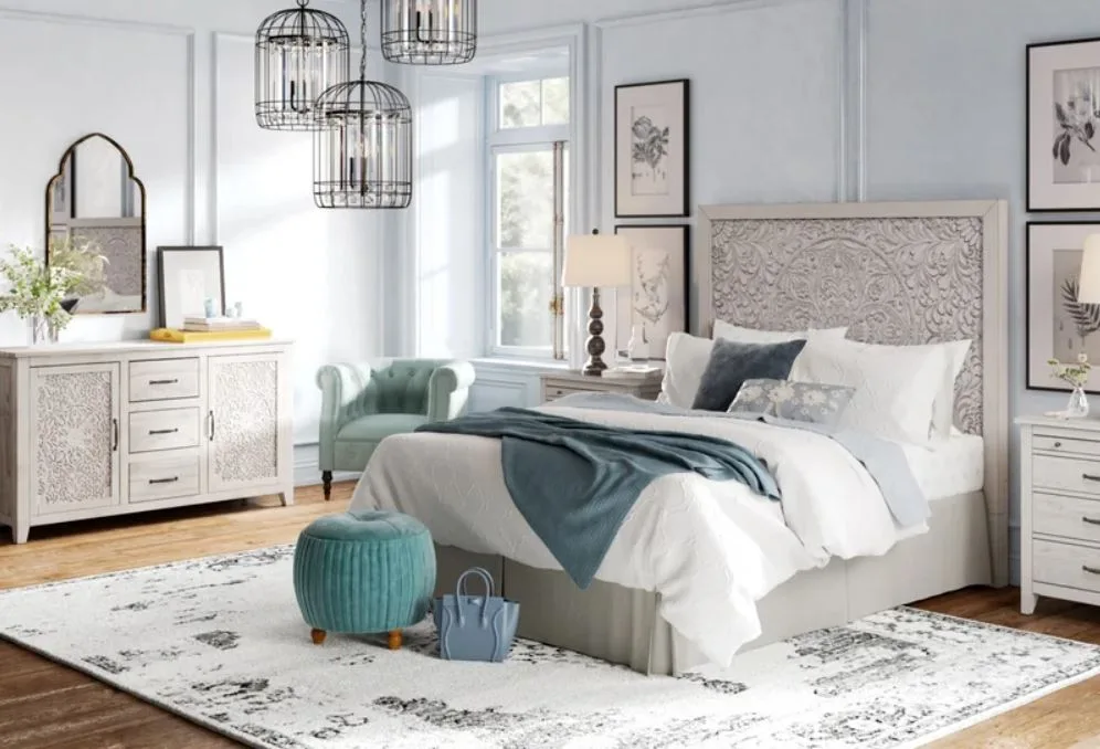 A bright bedroom featuring a bed with a carved grey headboard, a distressed white dresser, and teal accent furniture on a patterned area rug.