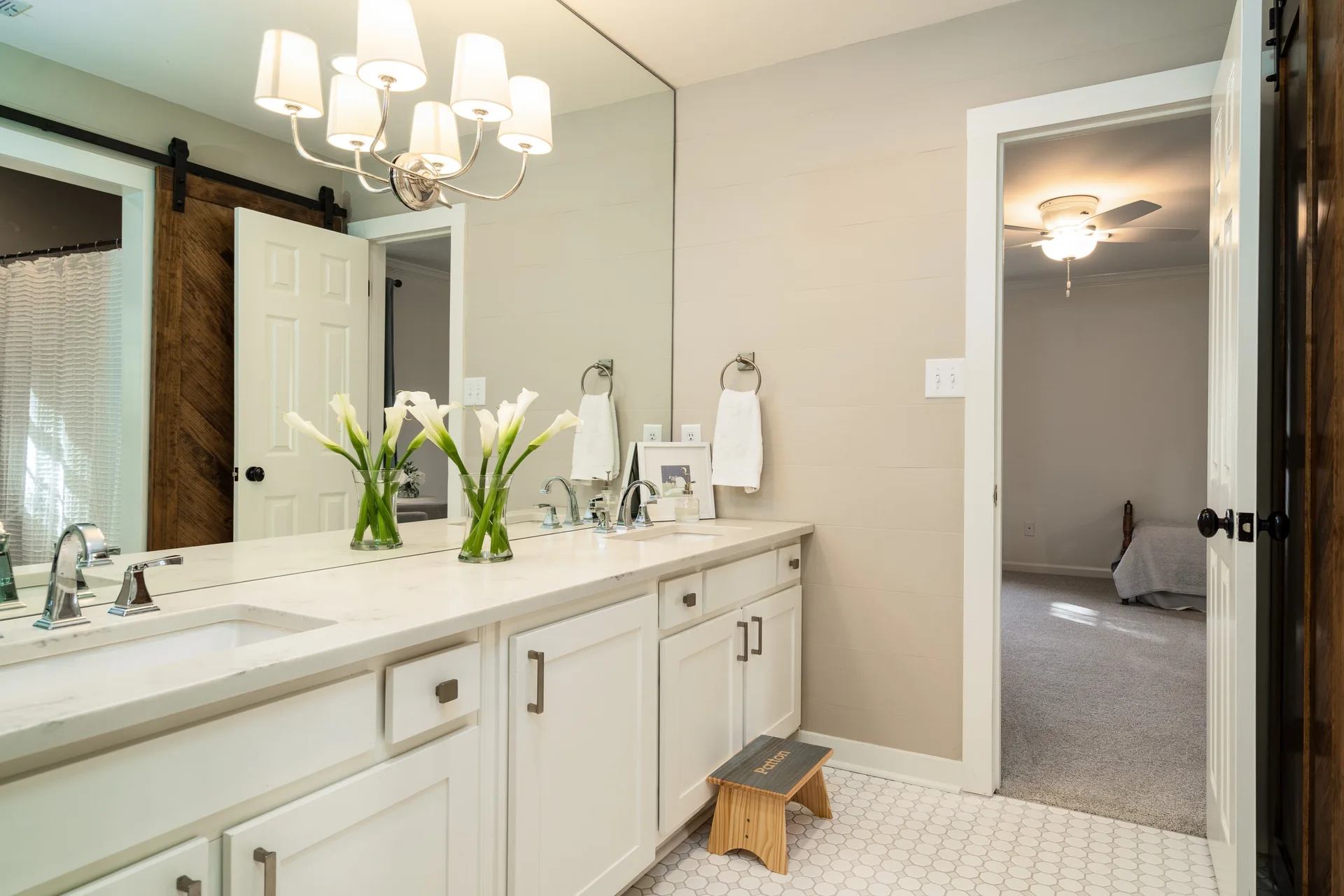 A bright bathroom features a long white double-sink vanity with a large mirror, beige horizontal plank walls, and small white hexagonal floor tiles.