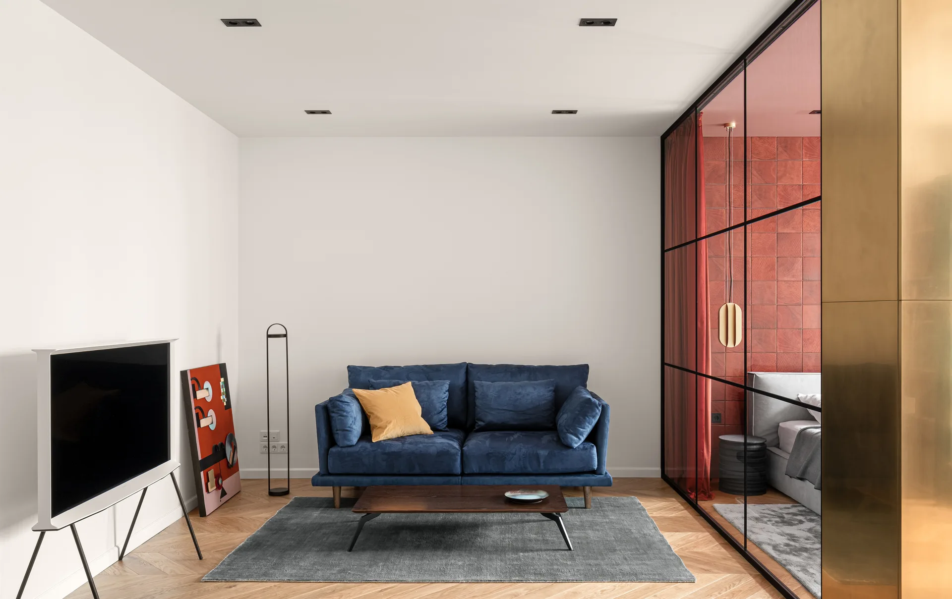 A living room features a blue velvet sofa on a grey rug, a dark wood coffee table, and a black-framed glass partition separating it from a bedroom.