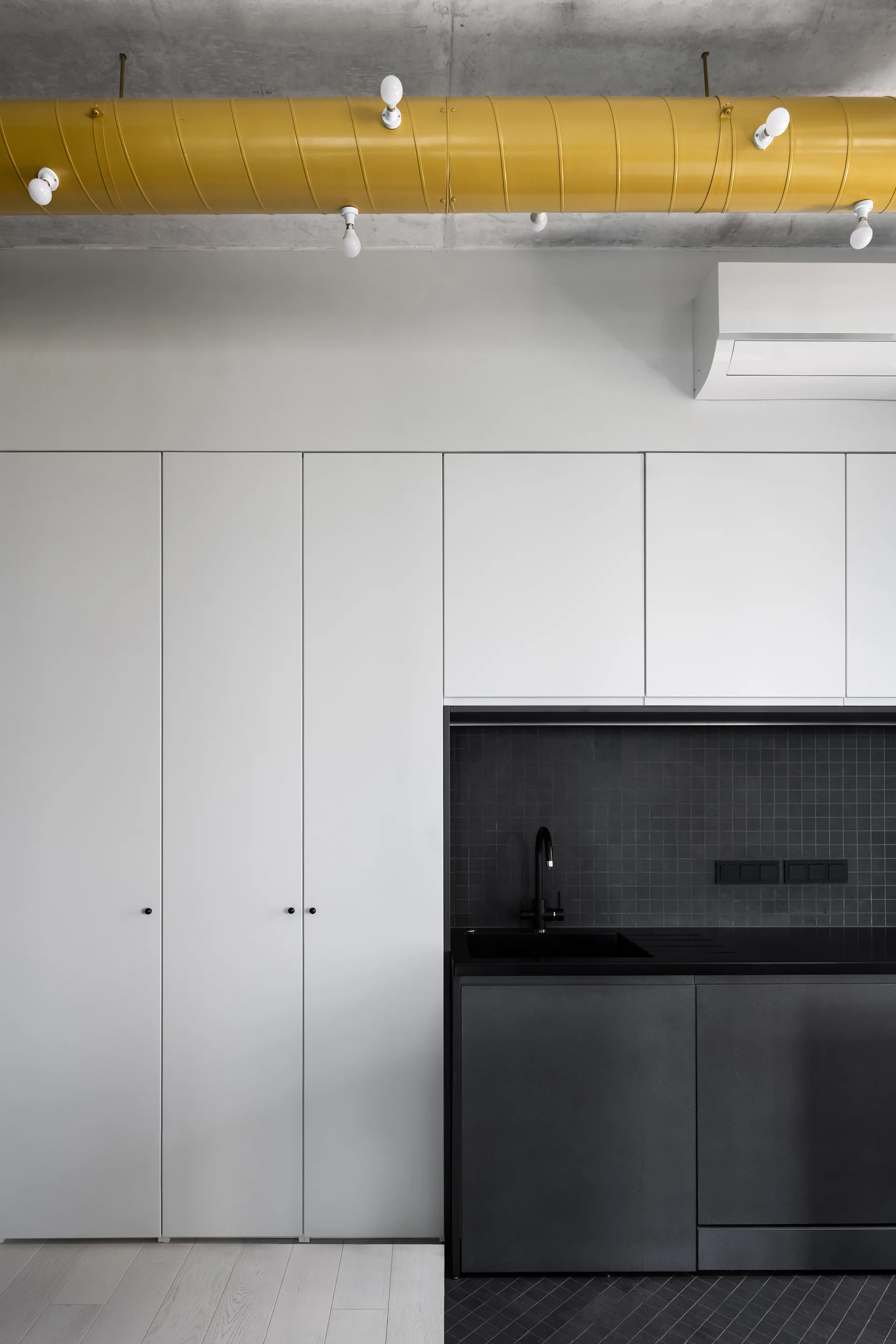 A minimalist kitchen features floor-to-ceiling white cabinets, a dark charcoal sink area with a grid tile backsplash, and an exposed yellow ceiling duct.