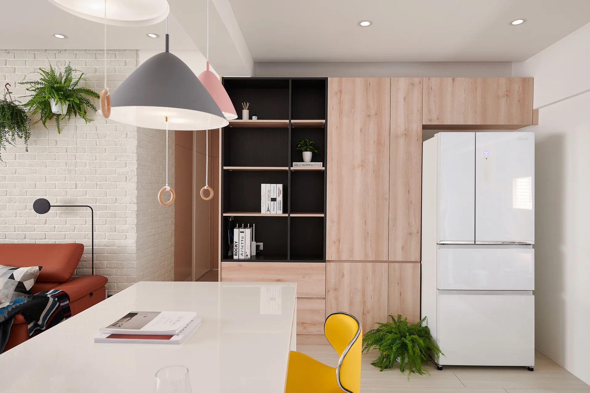 A modern dining area with a white table, a yellow chair, colorful pendant lights, light wood cabinets, and a white brick wall.