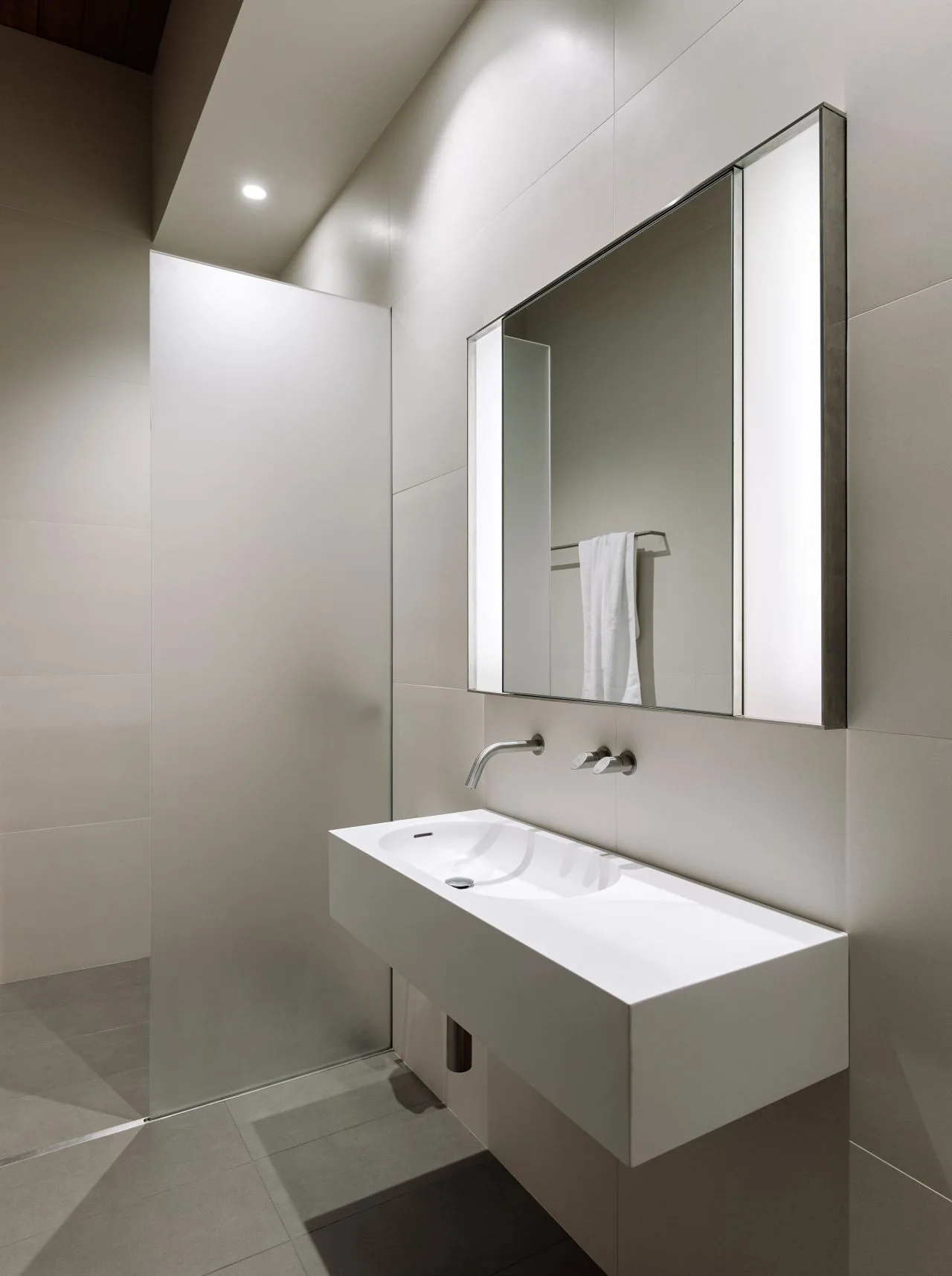 A minimalist bathroom features a white wall-mounted sink, a rectangular illuminated mirror, and a frosted glass partition next to beige wall tiles.