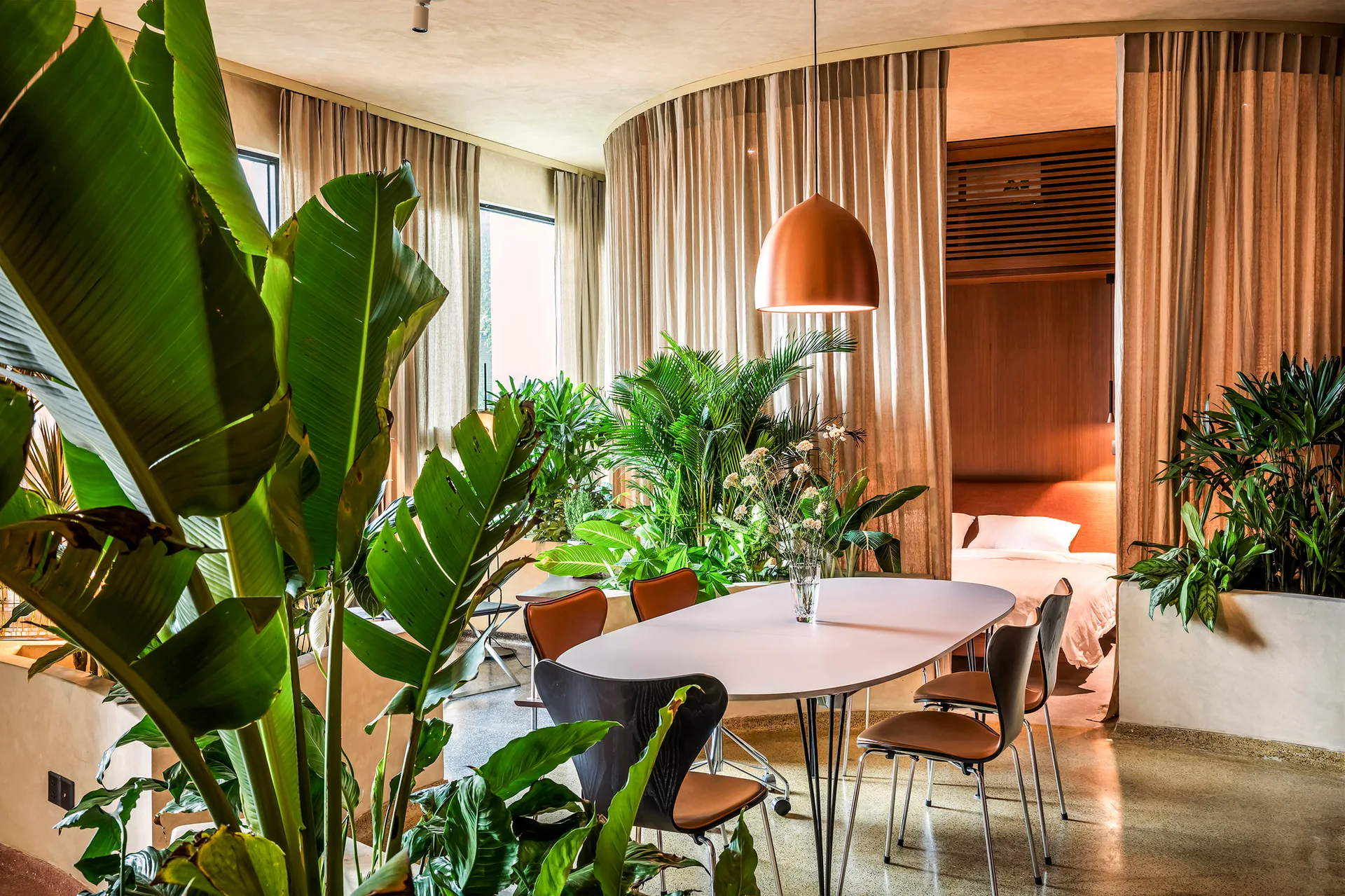 A modern dining room with an oval table, copper pendant light, and abundant green plants, separated from a bedroom by beige curved curtains.
