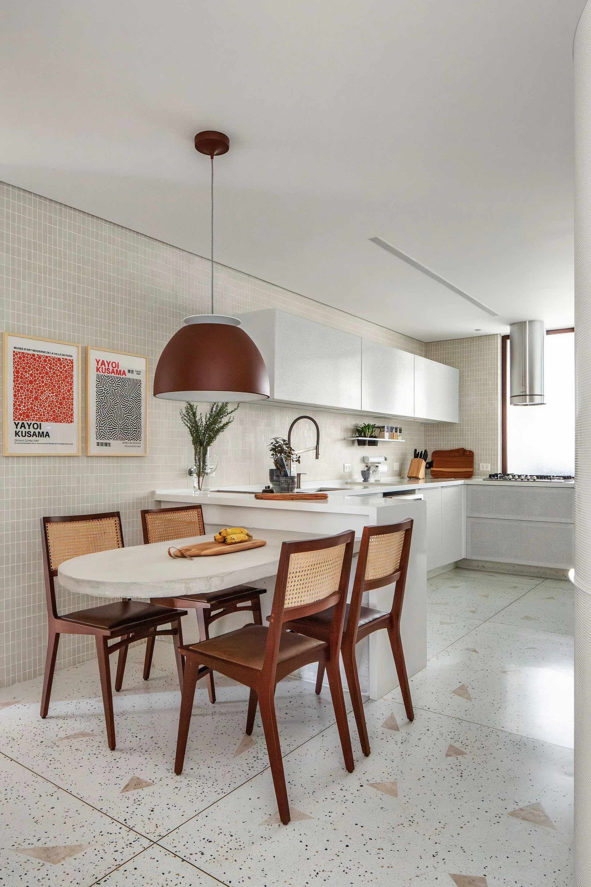 A modern white kitchen featuring an integrated oval dining table with cane-back chairs, terrazzo tile flooring, and a large brown dome pendant light.