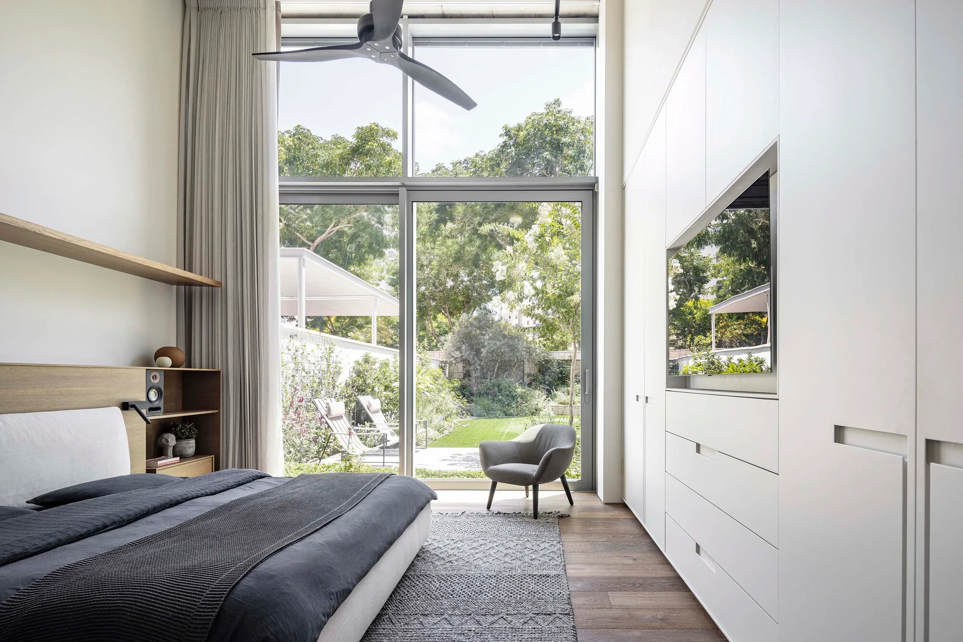 A modern bedroom with a low-profile bed, textured grey rug, built-in white wardrobes, and a large window looking out at a green garden.
