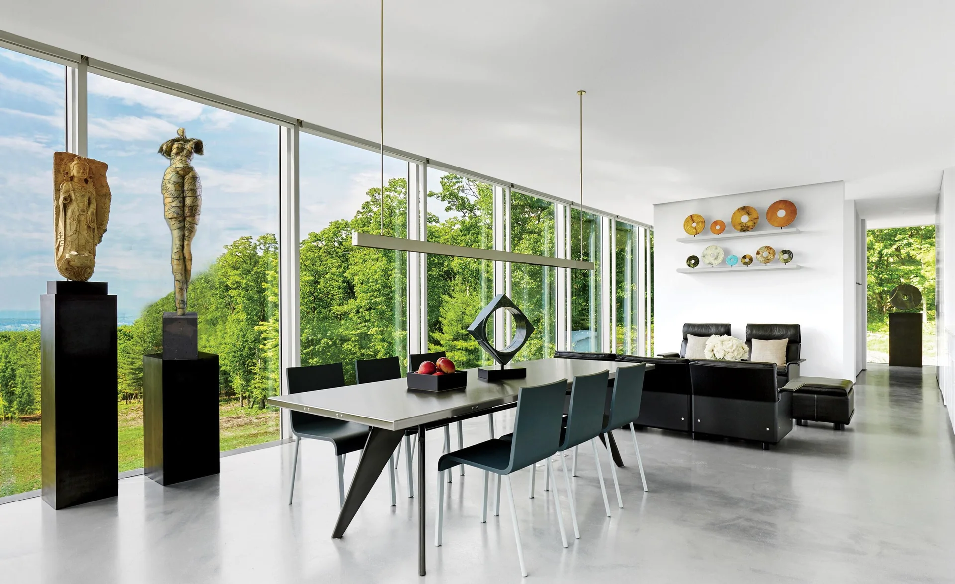 A modern dining room featuring a metal table with dark chairs, tall pedestals displaying sculptures, and large windows overlooking a dense forest.
