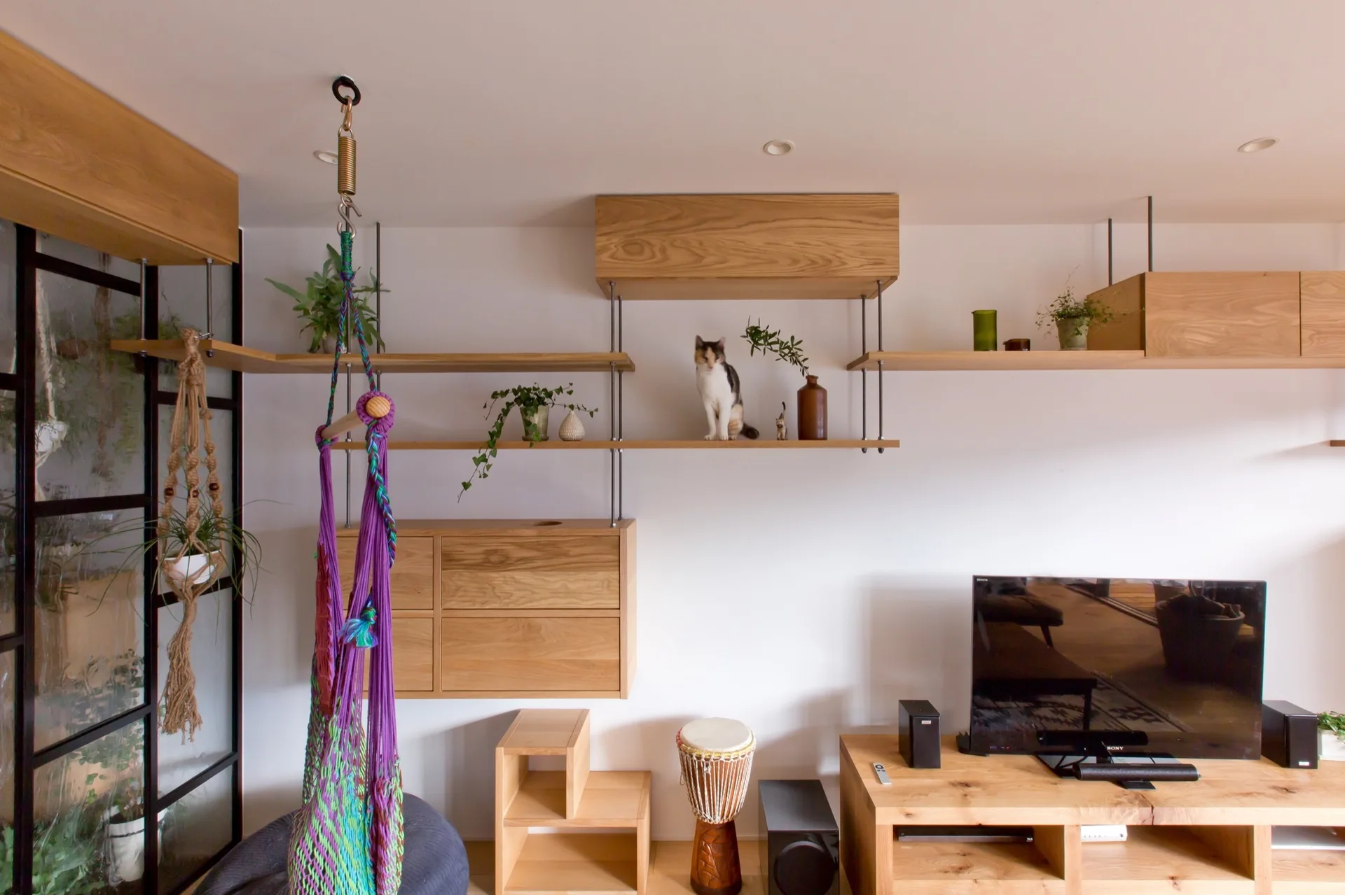 A living room features custom light wood shelving and cabinets mounted on a white wall next to a purple hanging chair and television console.