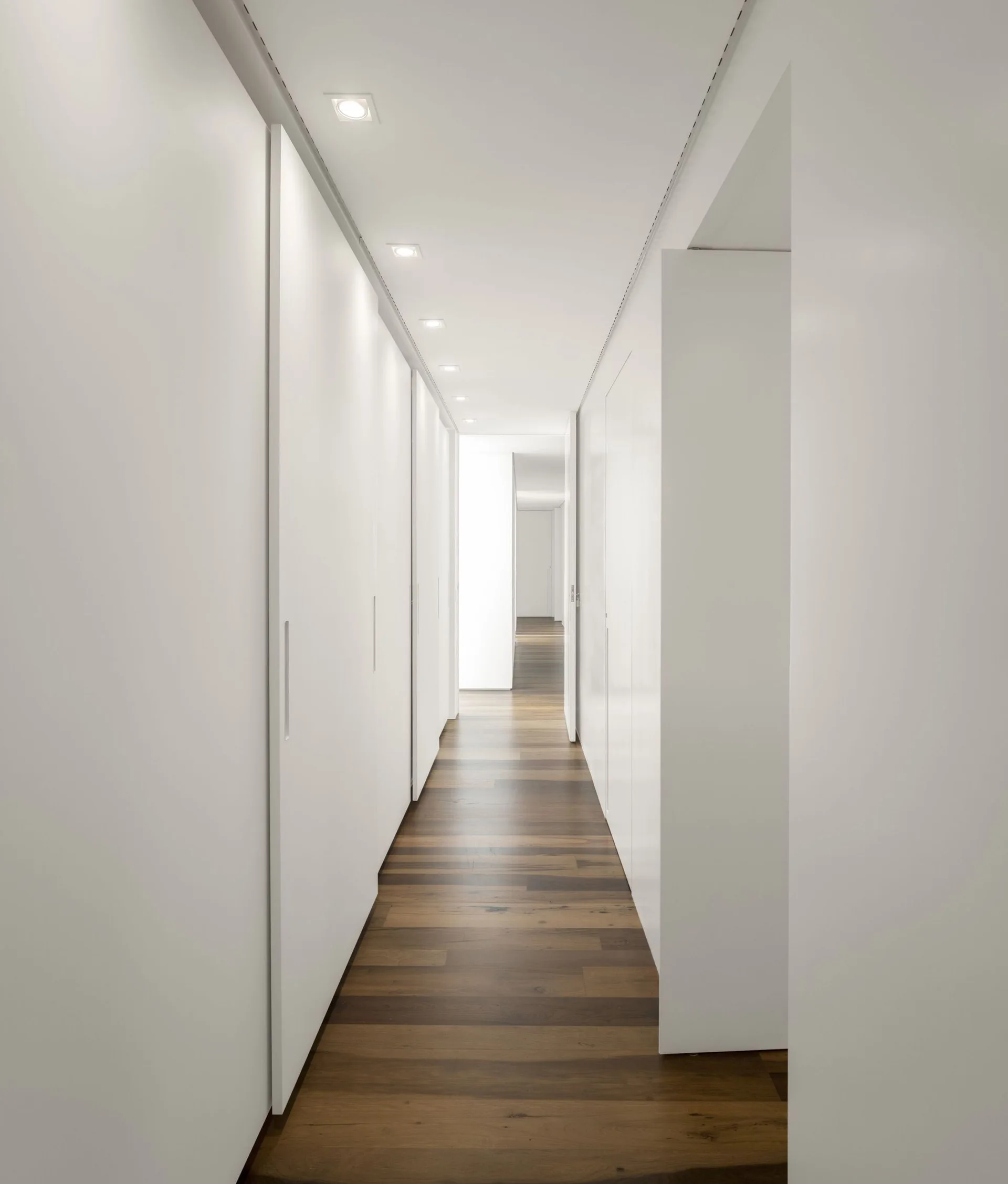 A long minimalist hallway features floor-to-ceiling white built-in cabinets, dark wood plank flooring, and square recessed lights in the ceiling.