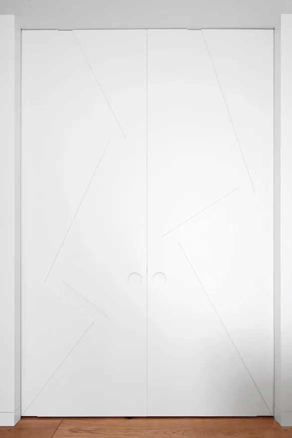 White minimalist double doors featuring incised geometric panel lines and circular integrated handle cutouts above wood flooring.