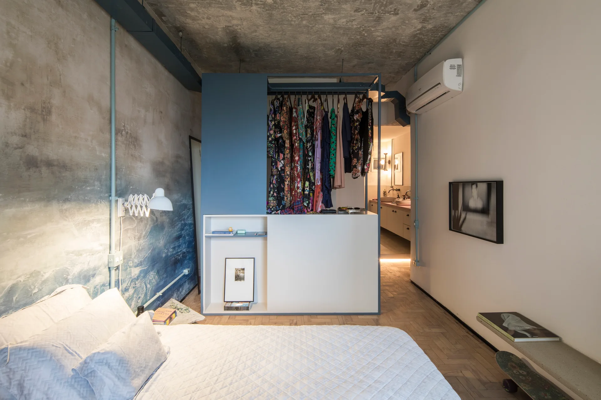 A modern industrial bedroom features a blue and white open closet room divider, a raw concrete ceiling, and a blue watercolor mural wall.