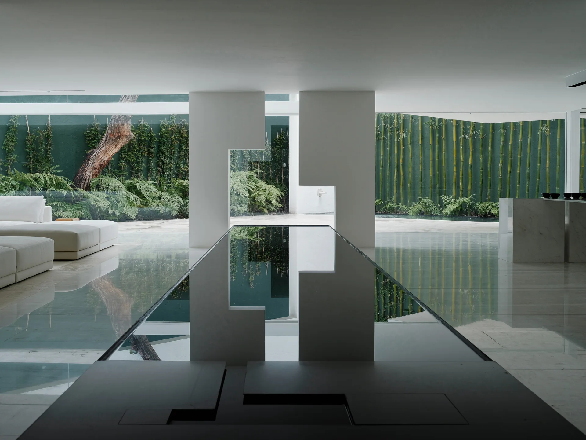 A minimalist living room features a white modular sofa and a long reflective table set before large windows overlooking a green courtyard garden.