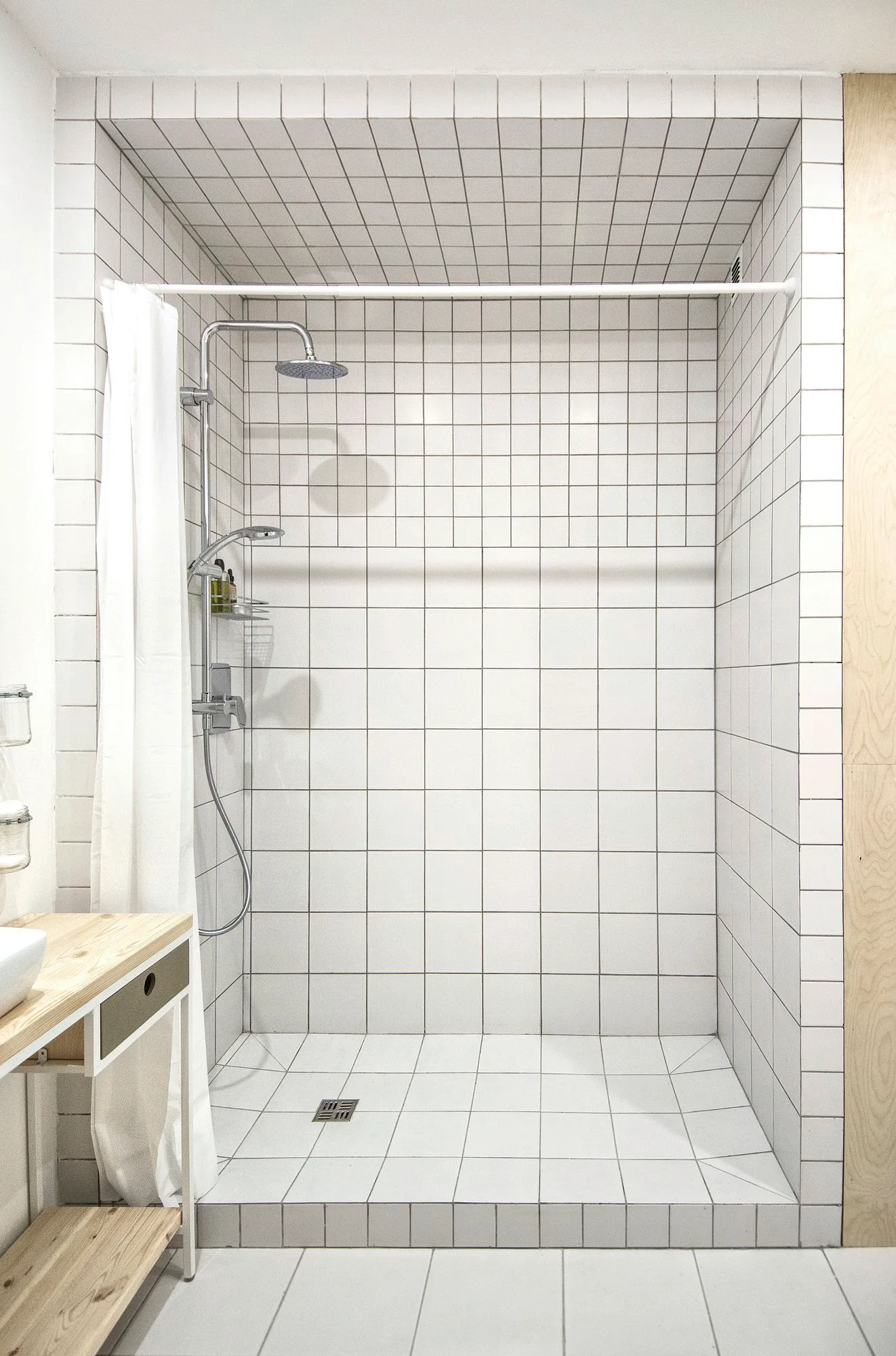 A walk-in shower lined with white square grid tiles features a chrome shower head, a white shower curtain, and an adjacent light wood vanity.