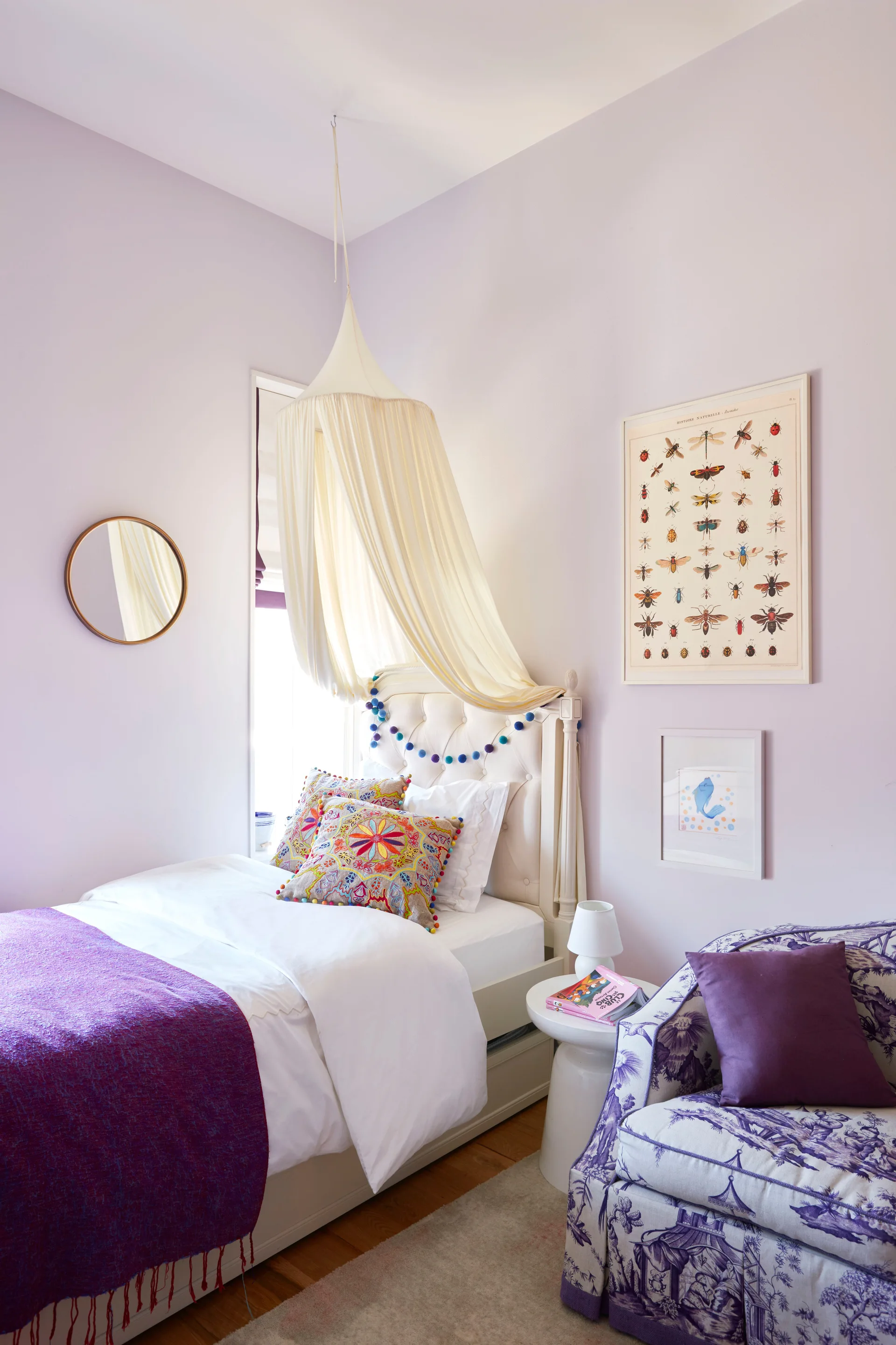 A lilac bedroom features a white bed draped with a fabric canopy, an upholstered armchair in a purple toile pattern, and framed wall art.