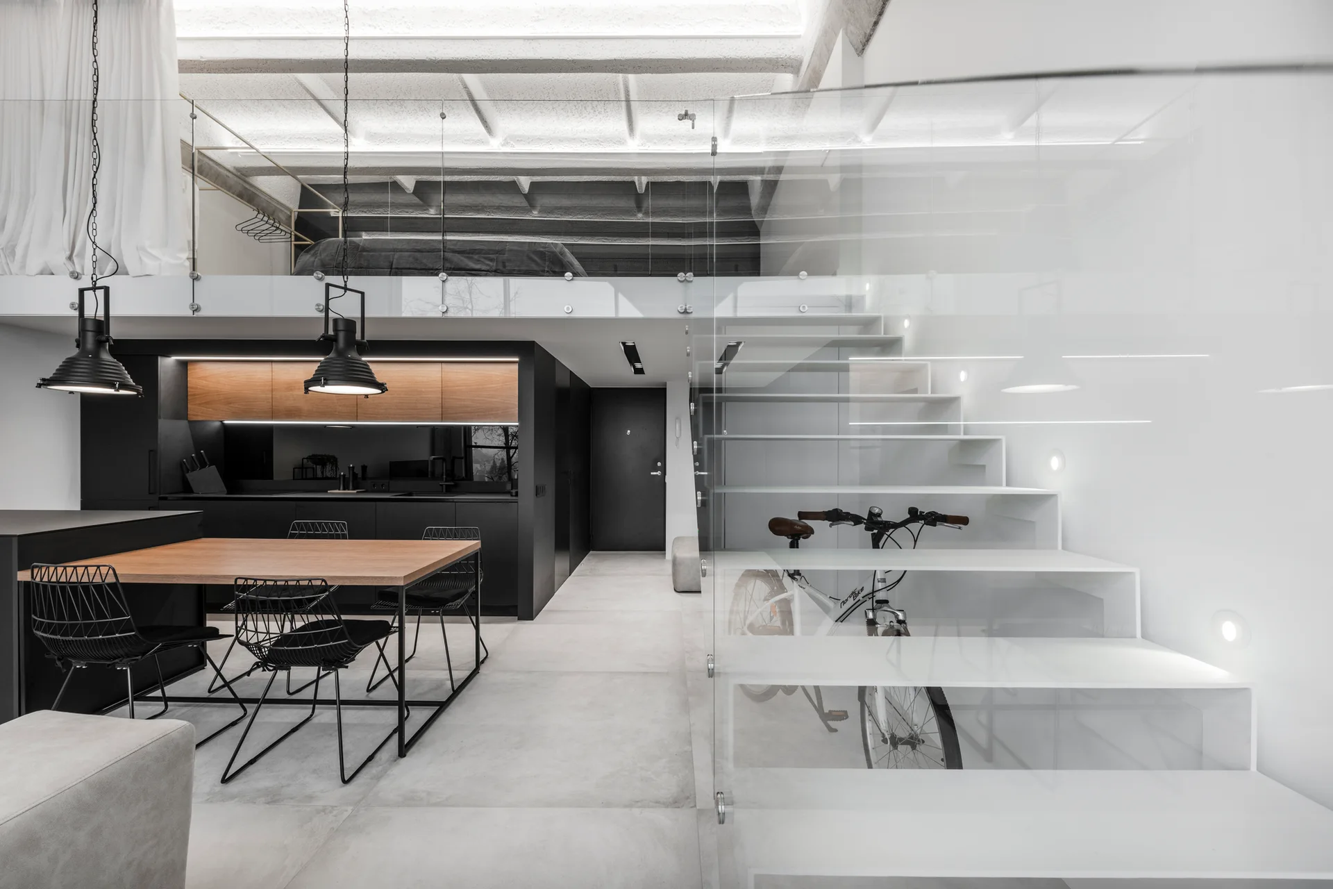 A modern minimalist loft features a floating white staircase with a glass railing, a black kitchen with wood accents, and an upper mezzanine bedroom.
