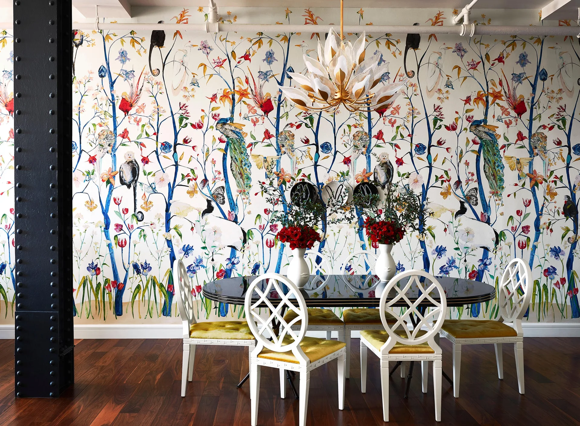 An eclectic dining room featuring a black oval table with white lattice-back chairs, a floral petal chandelier, and colorful botanical wallpaper.