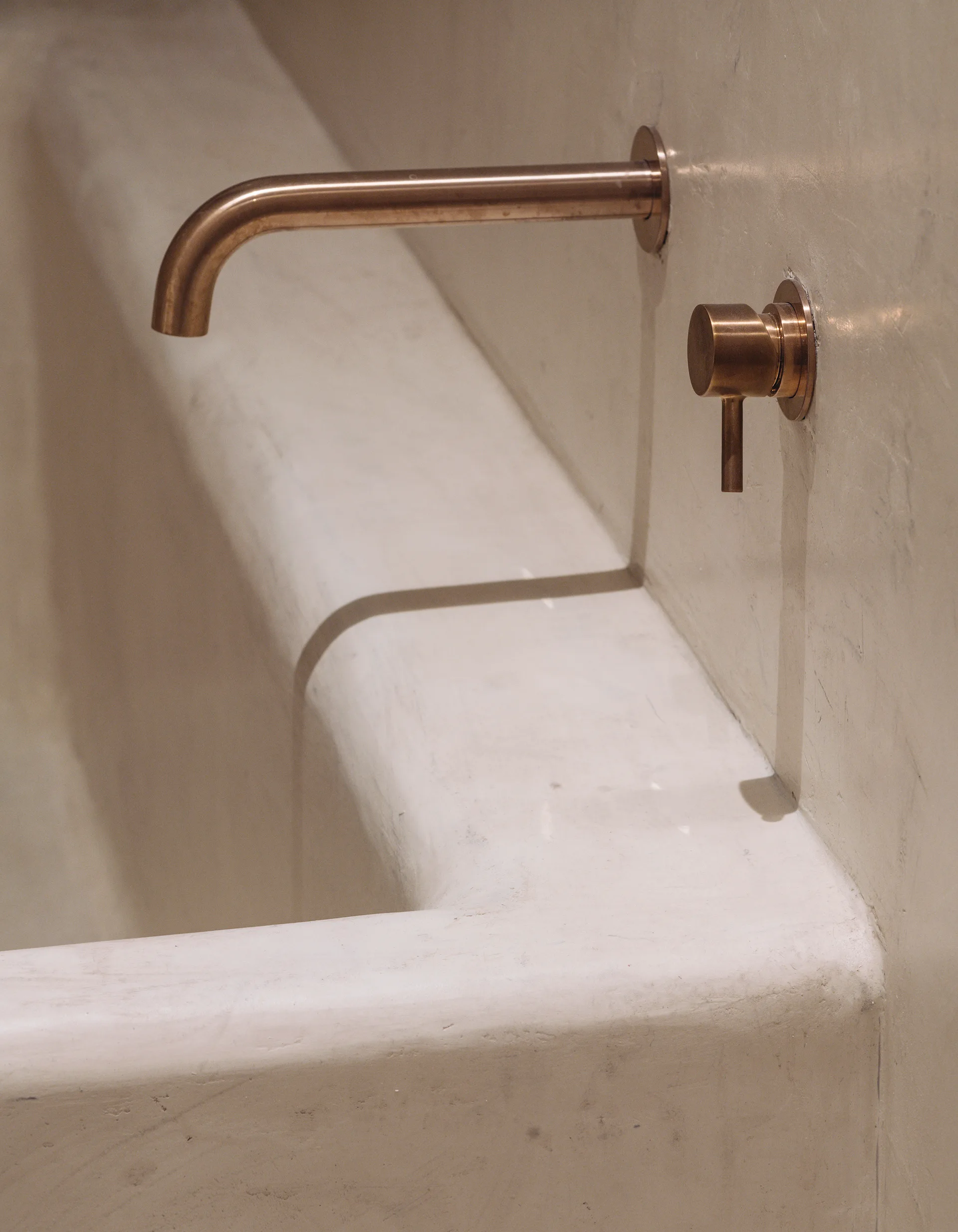 A detail shot of a wall-mounted bronze faucet and single-lever handle installed against a seamless off-white plaster bathtub wall.
