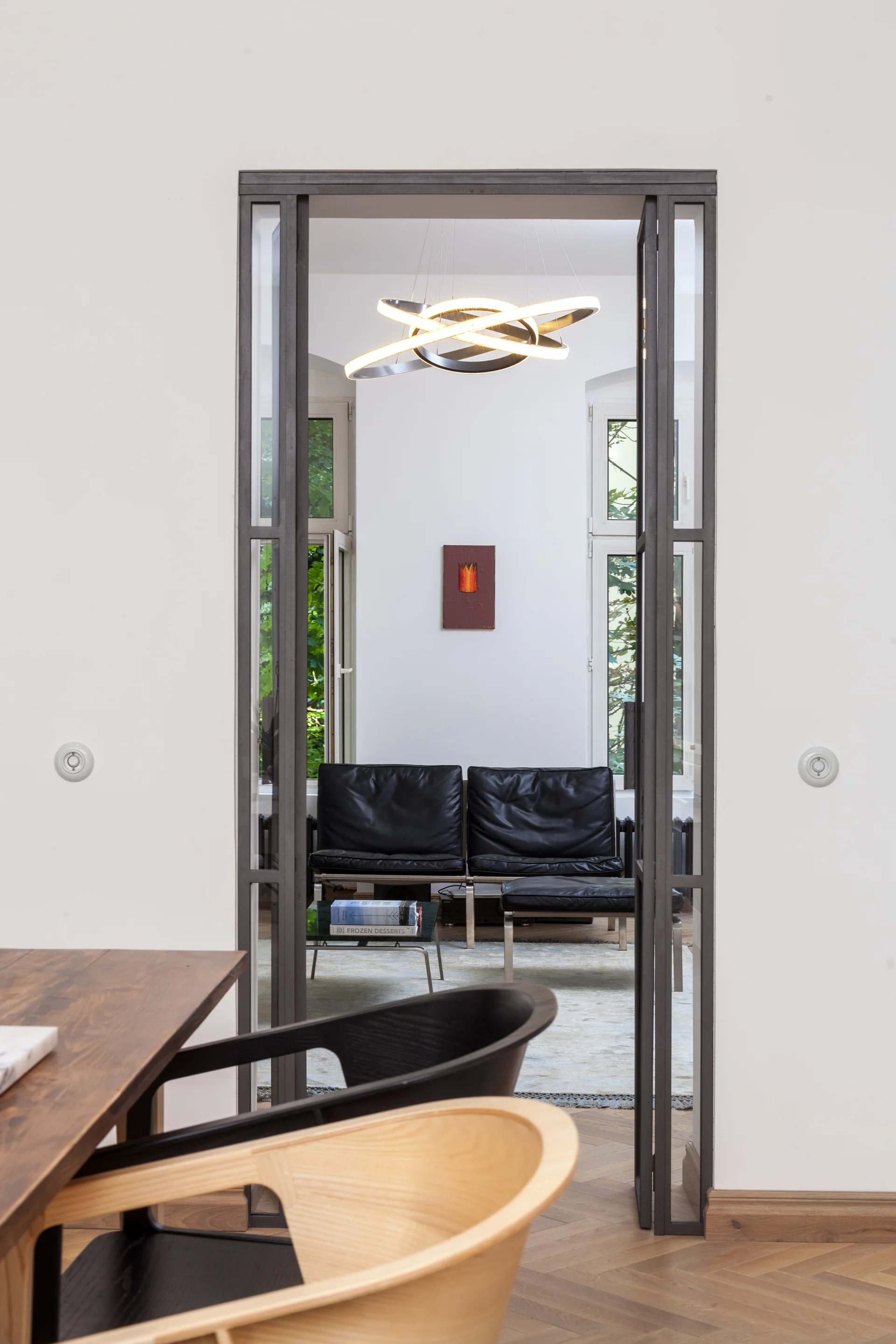 A view through a slim metal-framed doorway showing a living room with black leather armchairs, herringbone flooring, and a circular pendant light.
