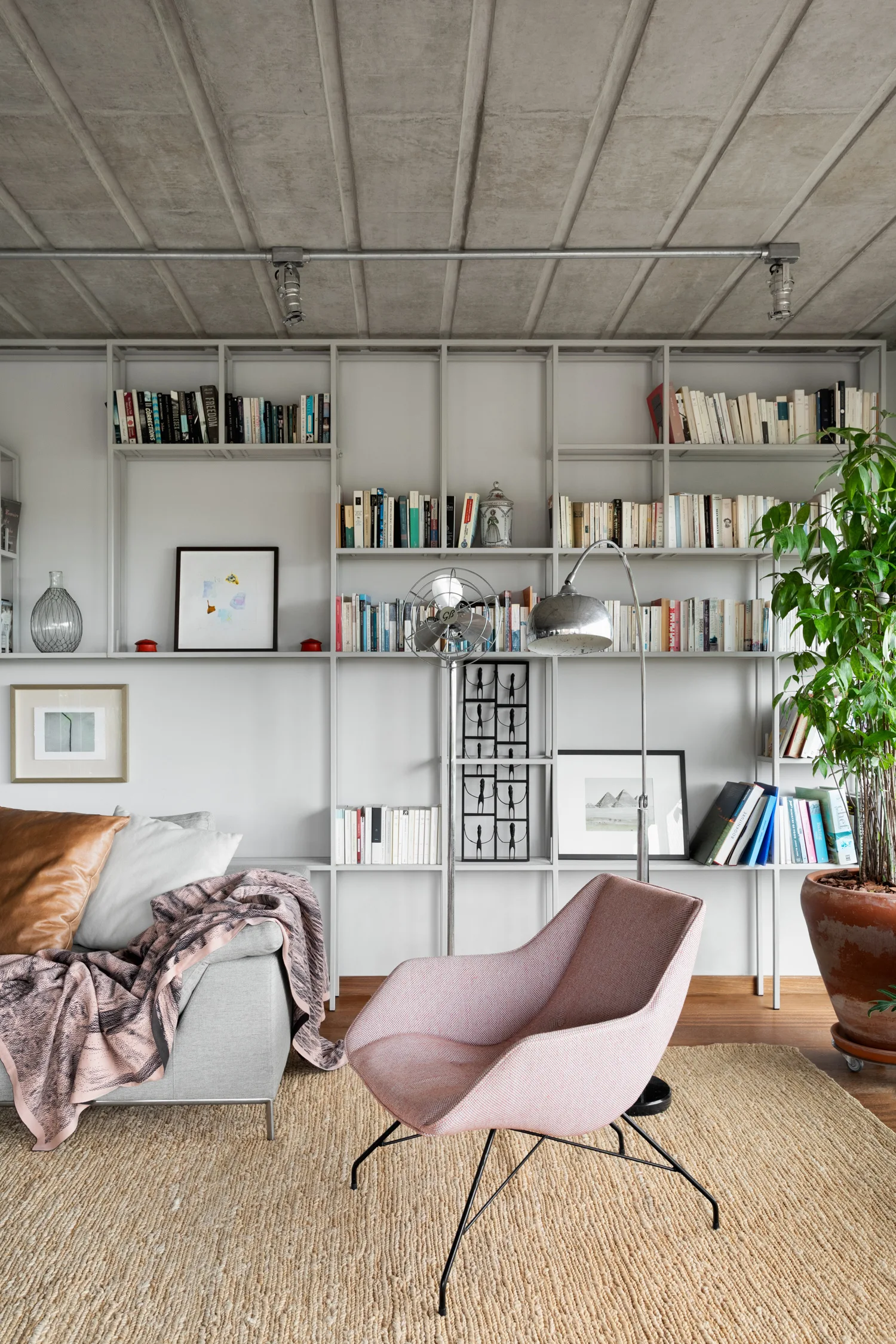 A living room features a pink armchair and grey sofa on a woven rug beneath an exposed concrete ceiling with a metal grid shelving unit.