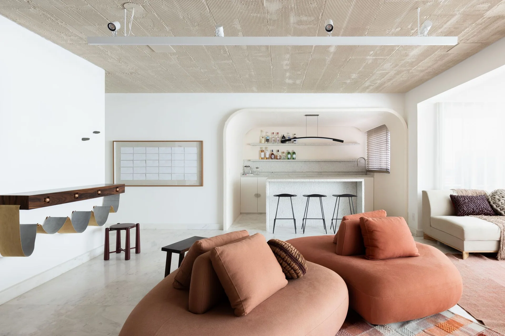 A minimalist living room features a curved terracotta modular sofa, a textured ribbed ceiling, and an arched alcove with a stone bar counter and stools.