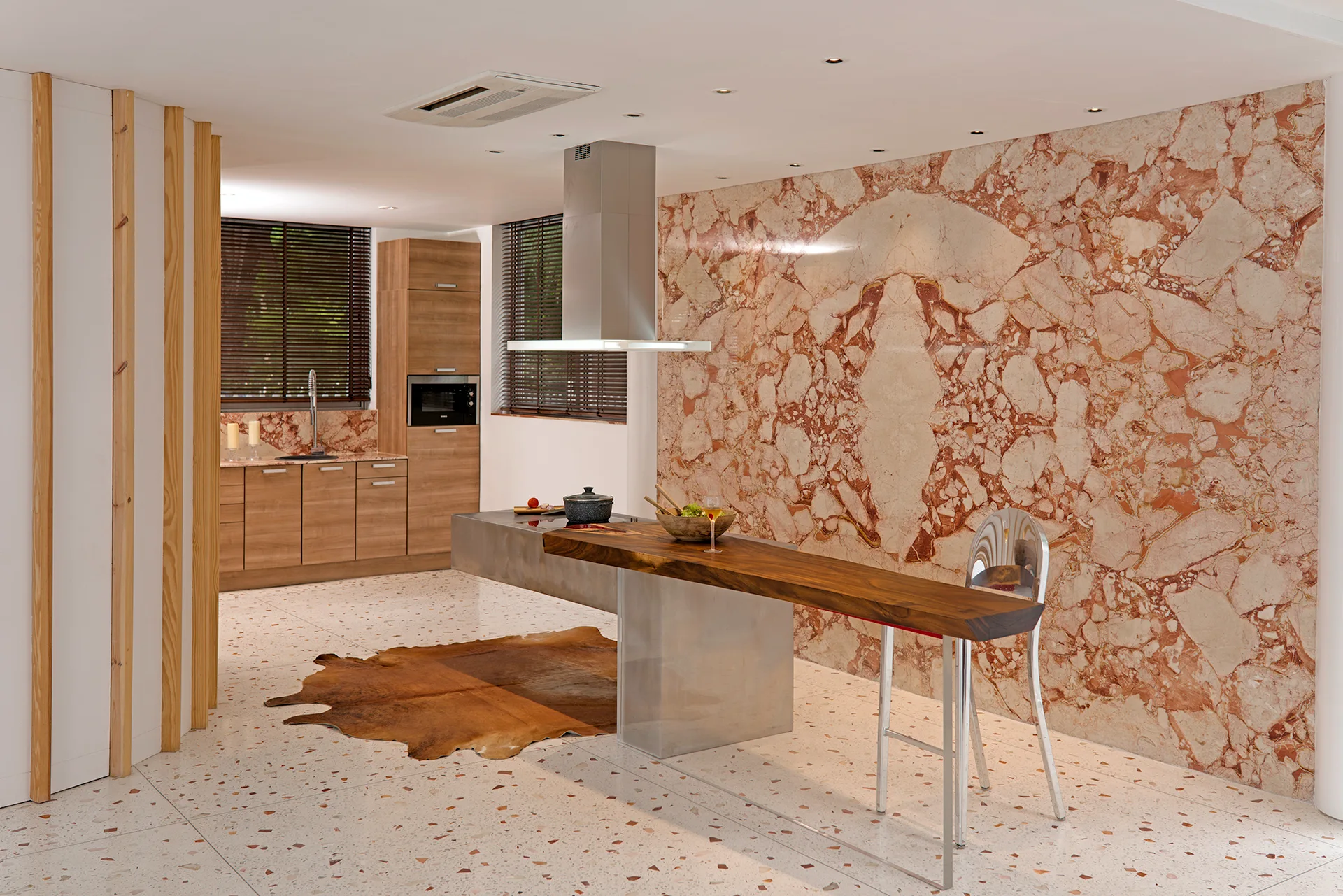A modern kitchen featuring a prominent pink marble accent wall, terrazzo flooring, and a stainless steel island with an attached wood bar top.