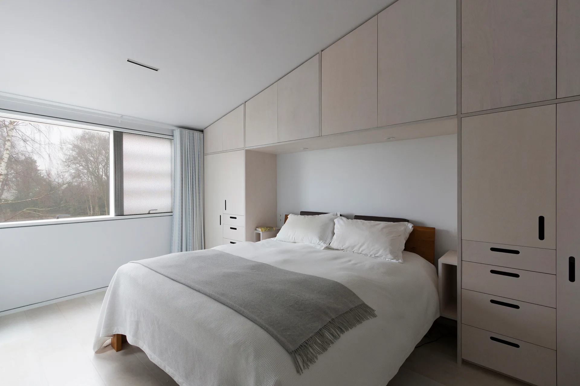 A modern bedroom with light wood built-in wardrobes framing a bed with white linens and a grey throw blanket under a sloped ceiling.