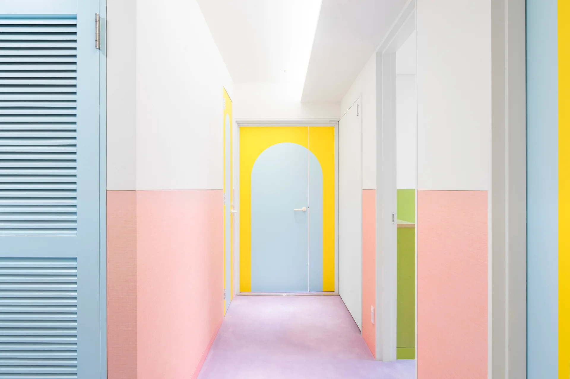 A brightly colored hallway features pink wall panels, a lavender floor, and a light blue door framed by a painted yellow arch.
