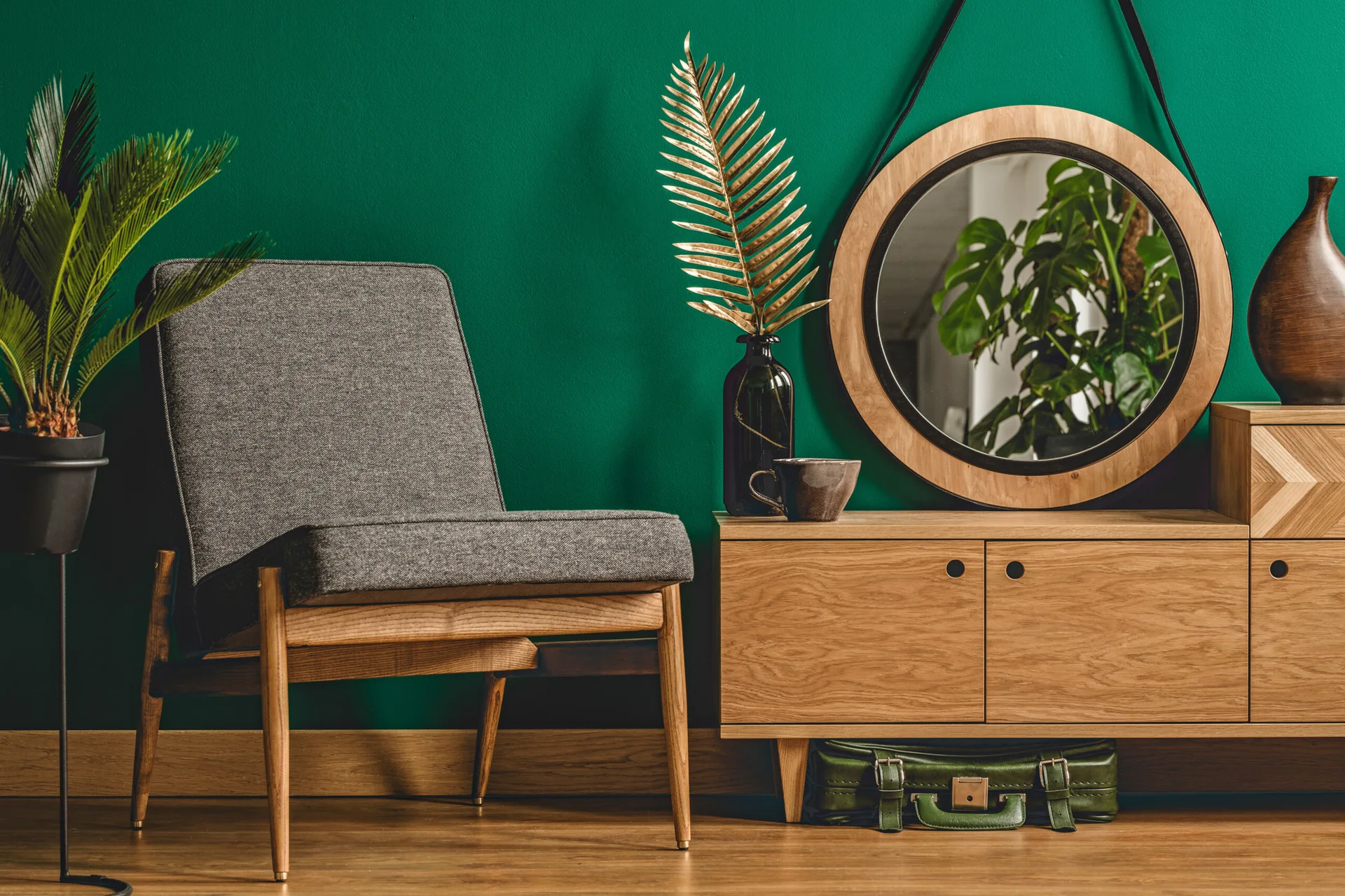 A grey fabric armchair sits next to a light wood sideboard against a deep green accent wall, topped with a round mirror and a gold leaf decor.