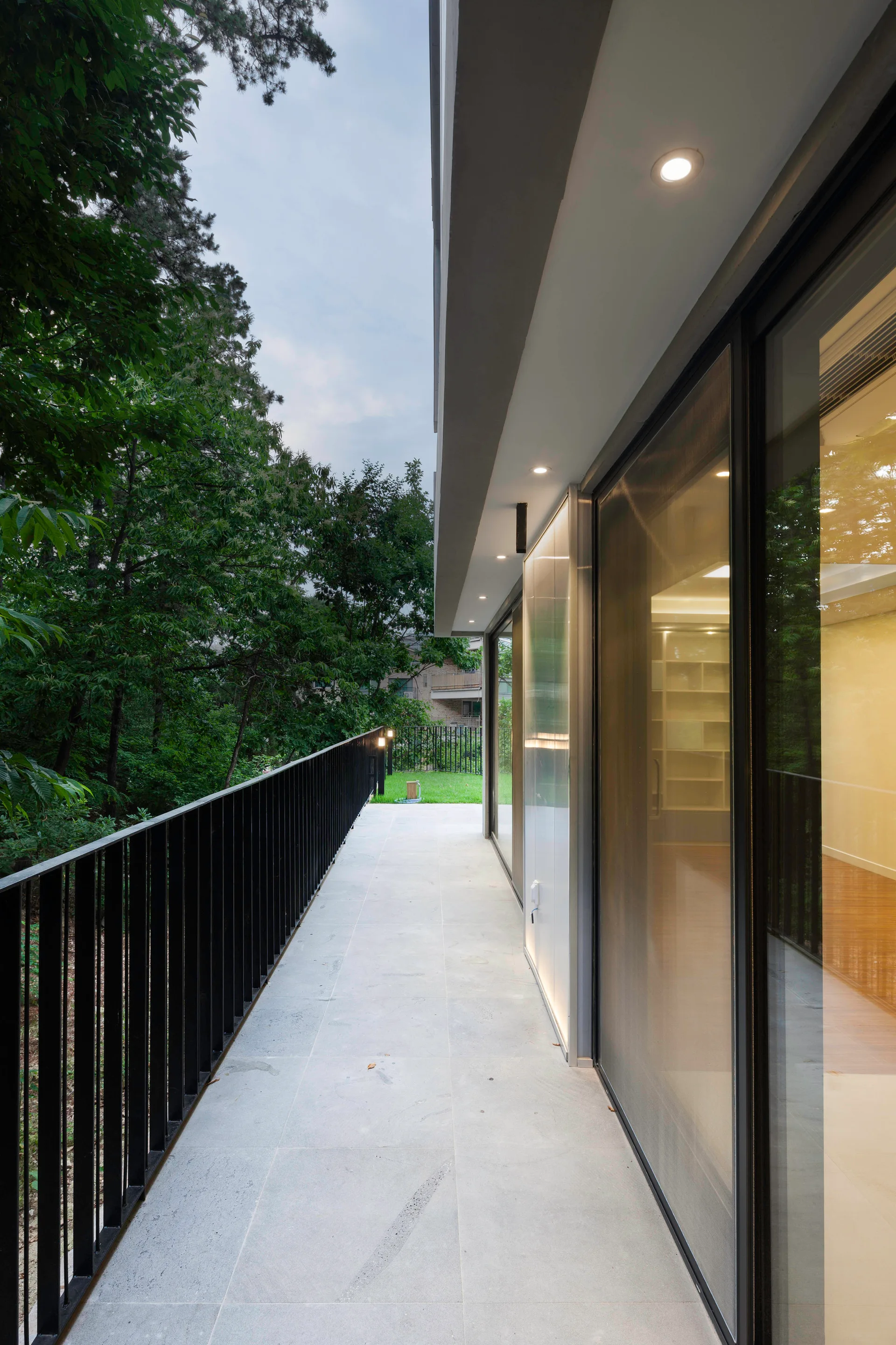 A modern balcony features light grey stone tile flooring, a black metal railing, and large glass sliding doors under a white ceiling with recessed lights.