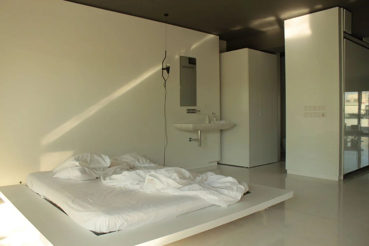 A minimalist white bedroom features a low platform bed with white bedding, a wall-mounted sink, and a contrasting dark ceiling.