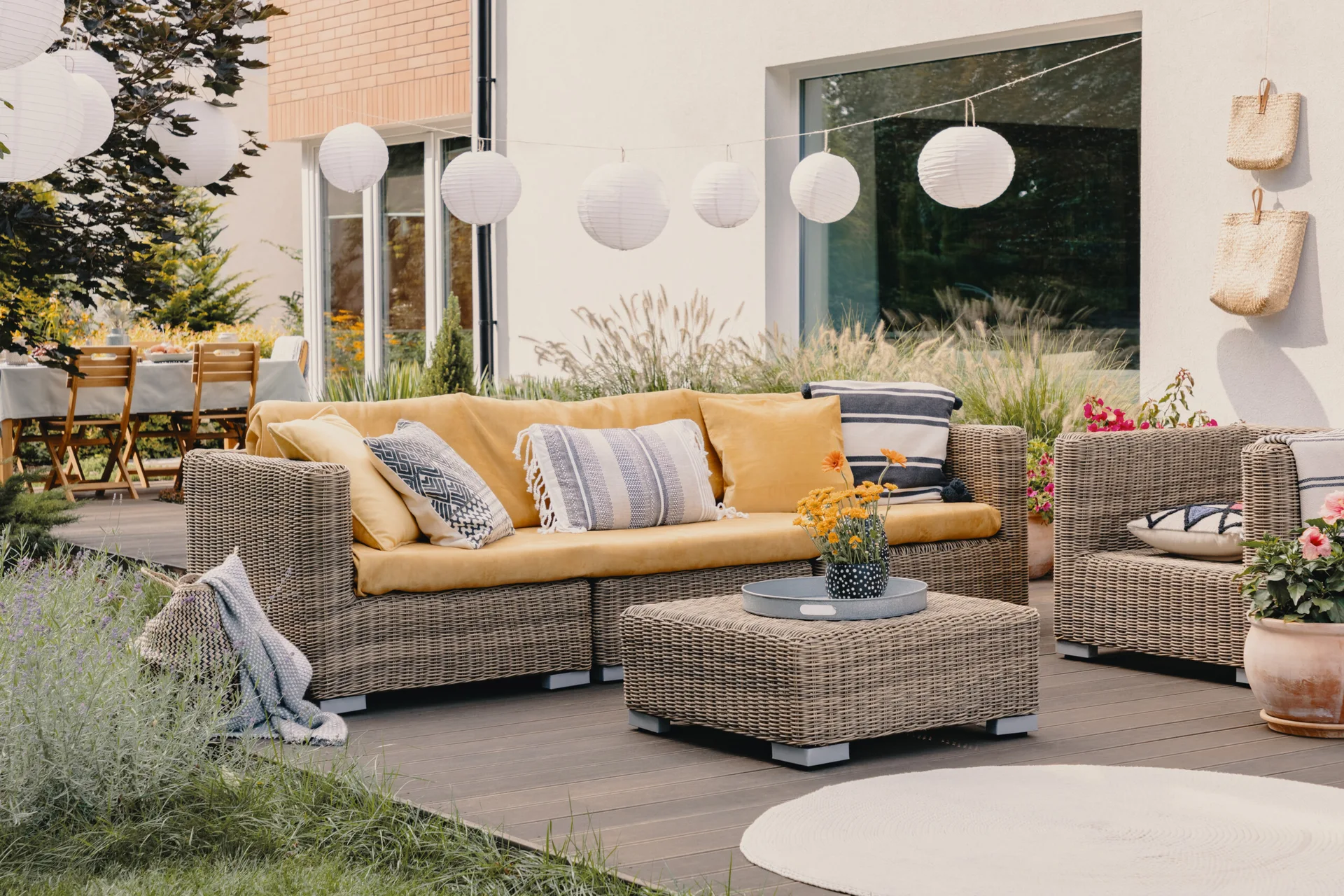 A wicker outdoor sofa with mustard yellow cushions and patterned pillows sits on a wood patio deck under suspended white paper globe lanterns.