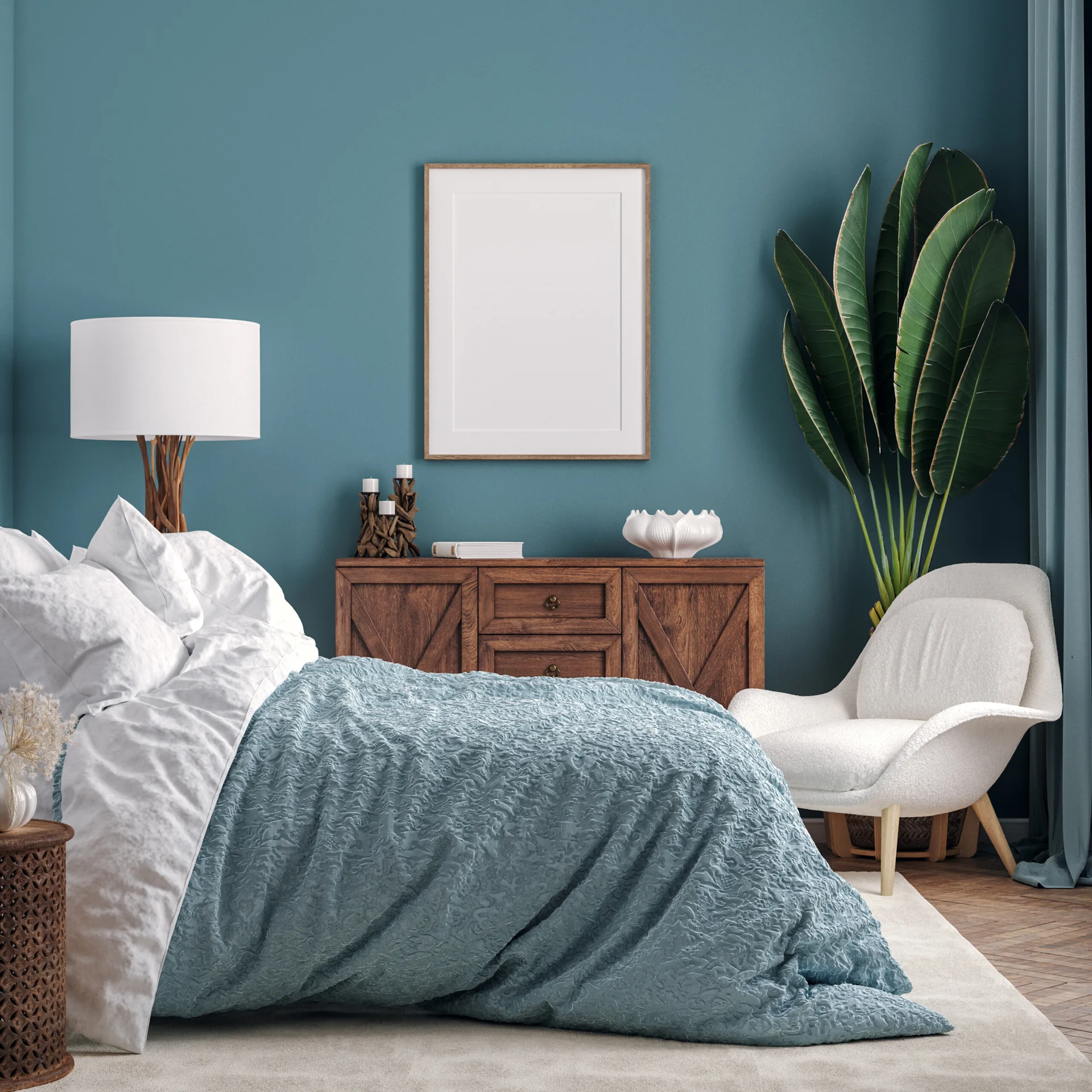 A bedroom featuring a textured blue bedspread, a wooden sideboard, and a white upholstered accent chair set against a deep teal wall.
