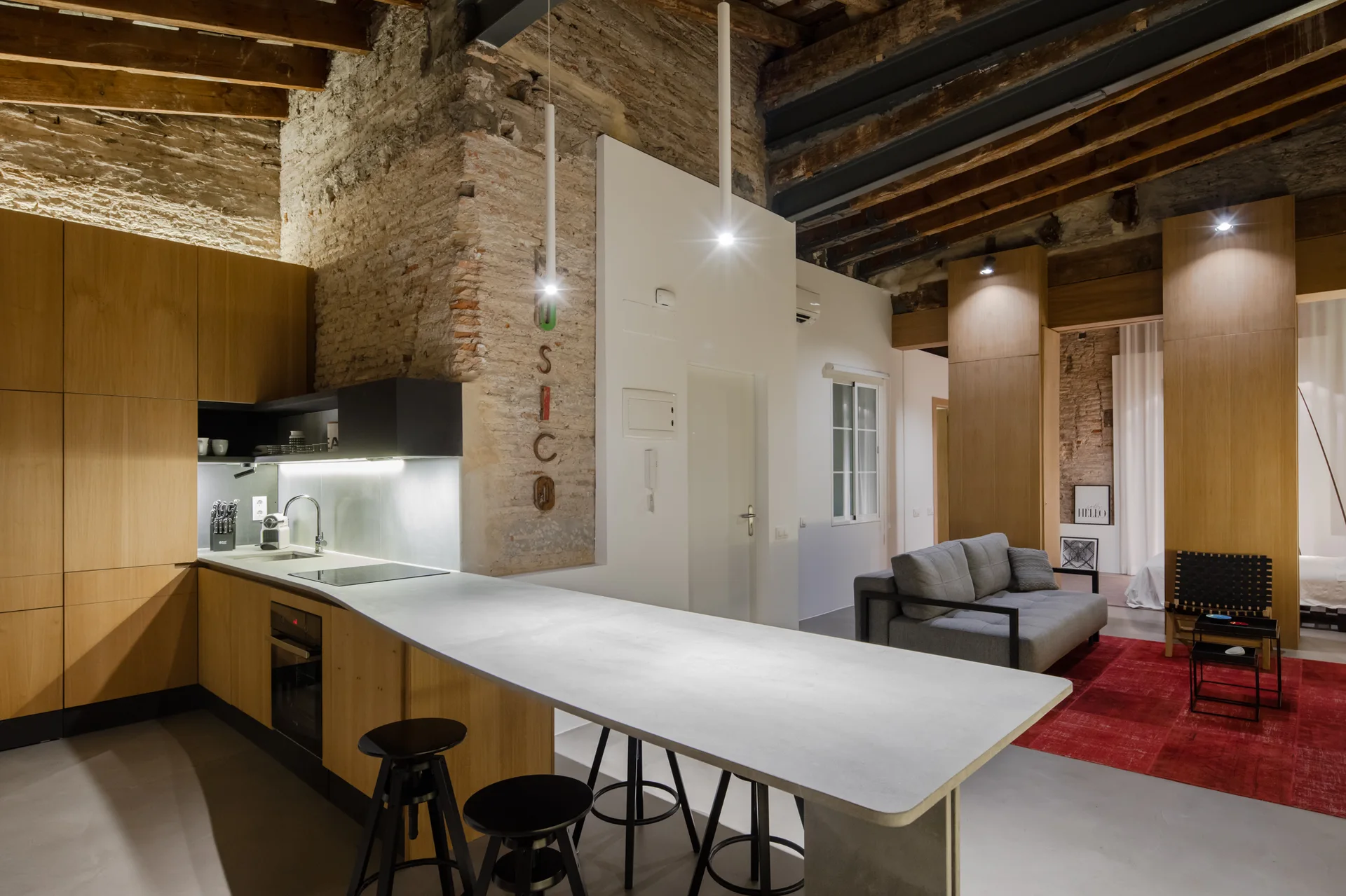 An industrial loft kitchen features flat-front wood cabinets, a long light grey island with black bar stools, and an exposed brick accent wall.
