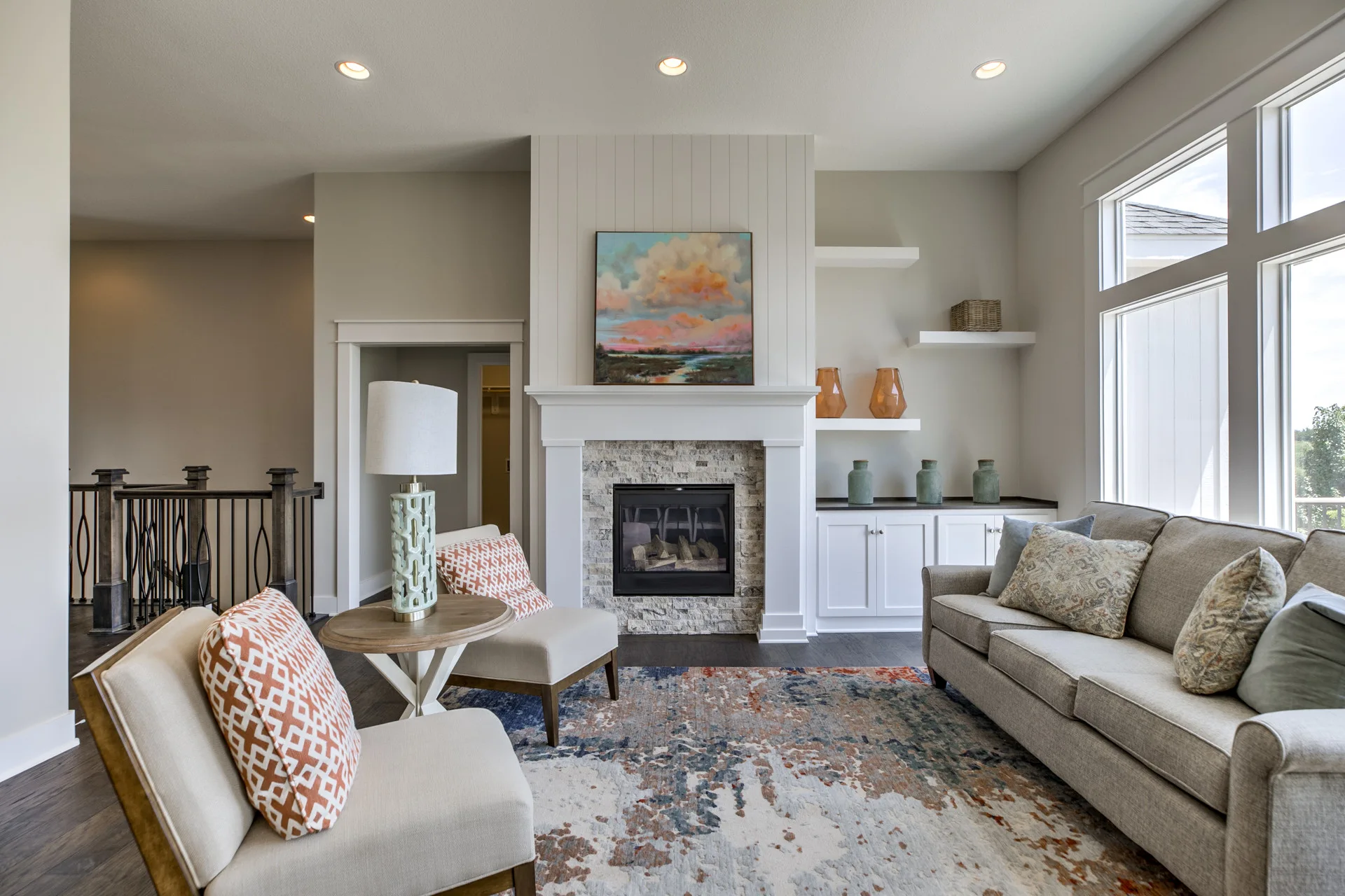 A transitional living room featuring a stacked stone fireplace with a white mantel, white built-in shelving, a gray sofa, and two armchairs.
