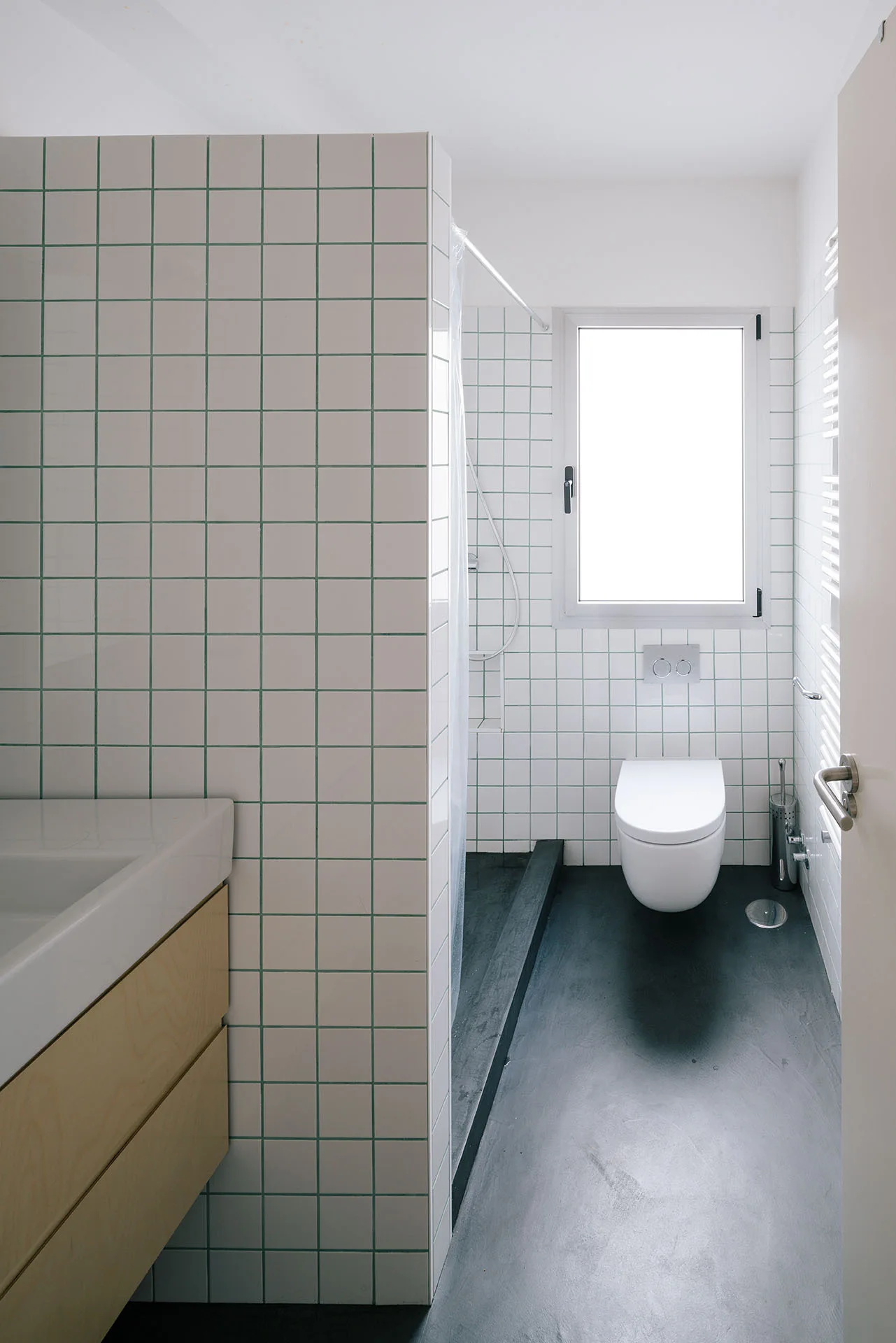 A minimalist bathroom features a white-tiled grid partition wall, a light wood floating vanity, a wall-mounted toilet, and a dark grey floor.