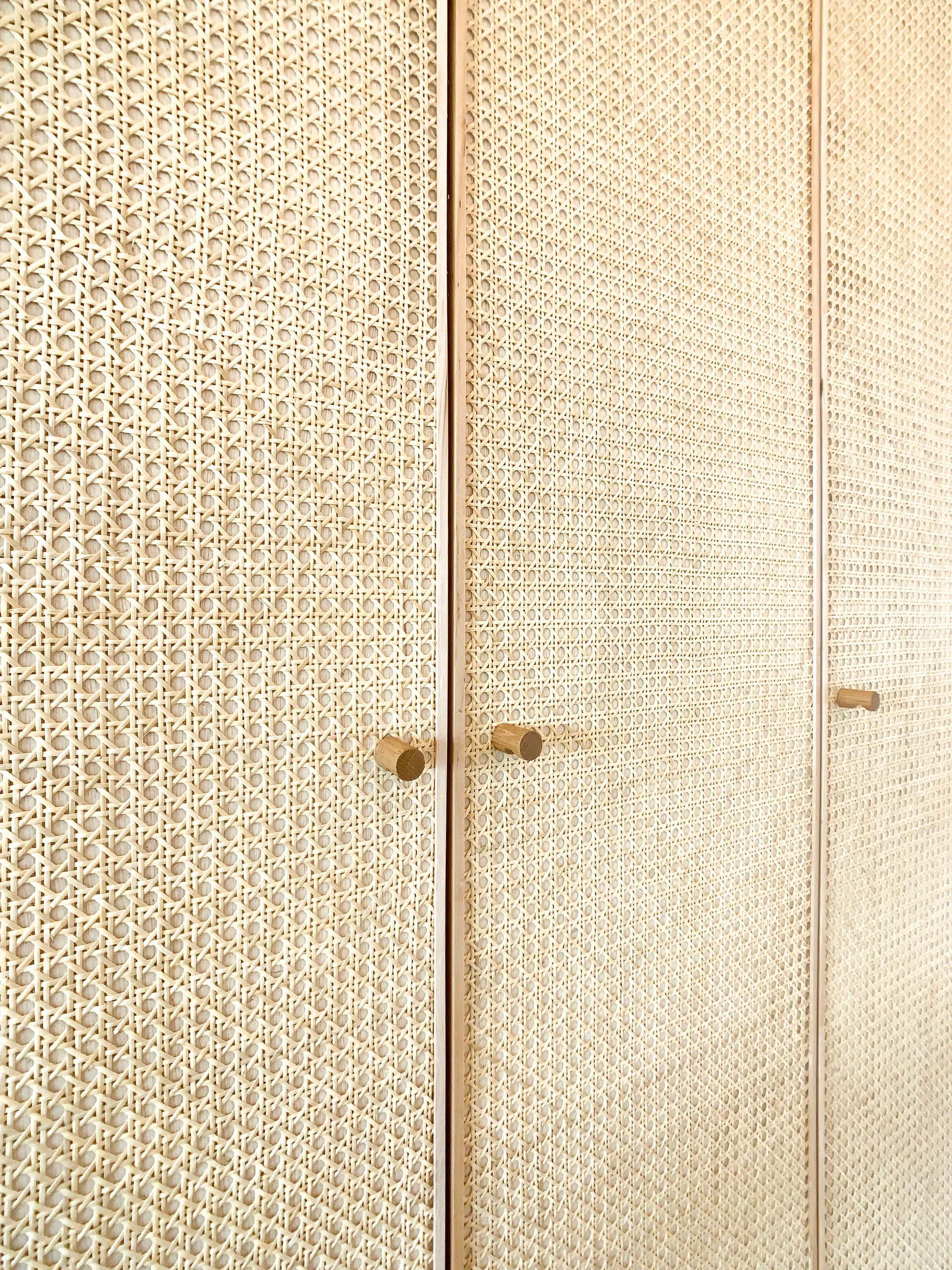 A close-up of three built-in cabinet doors covered in light beige woven cane mesh and fitted with simple, cylindrical wooden peg handles.