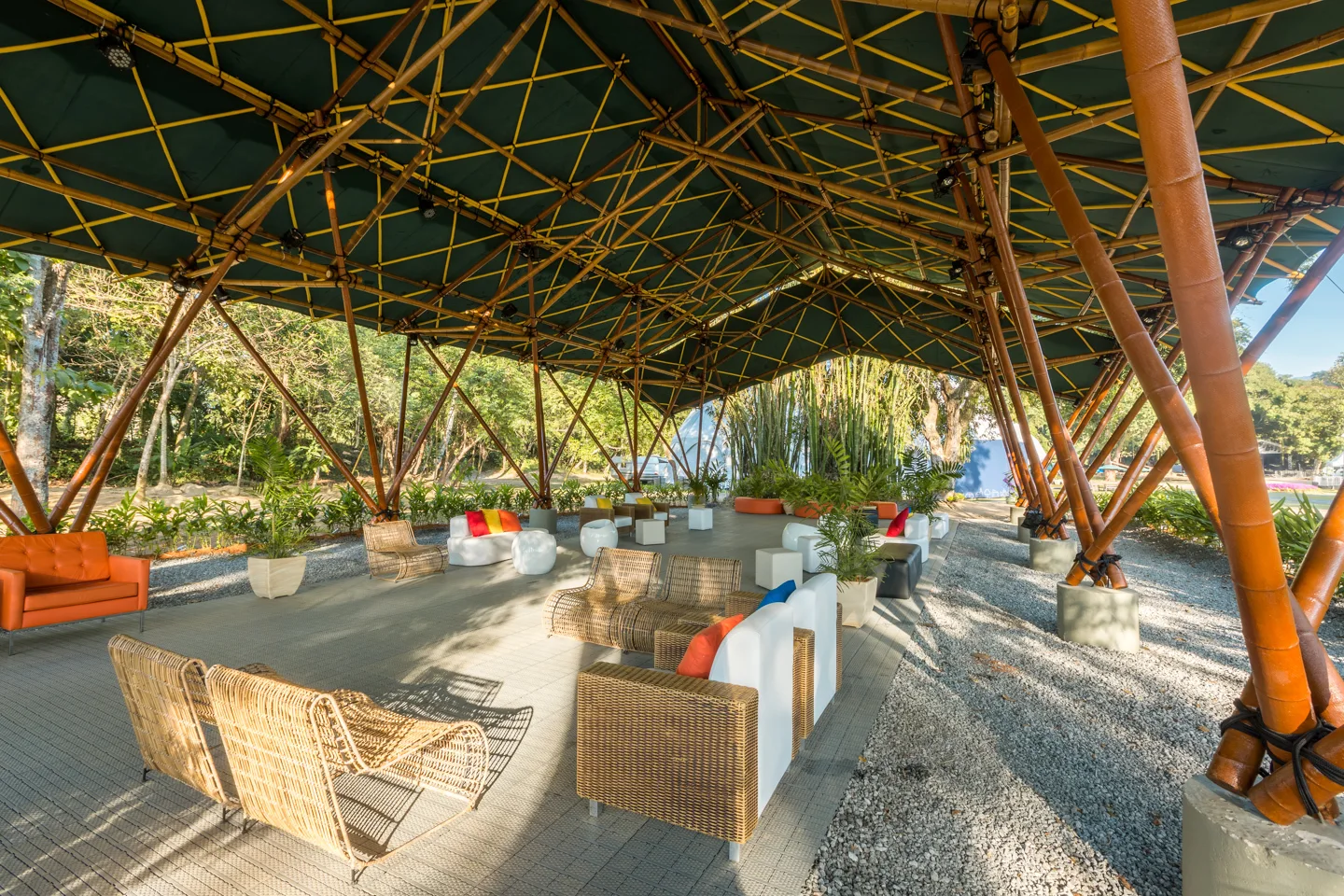 A spacious open-air patio under a large geometric bamboo pavilion features modern wicker armchairs and white sofas arranged on tiled and gravel ground.