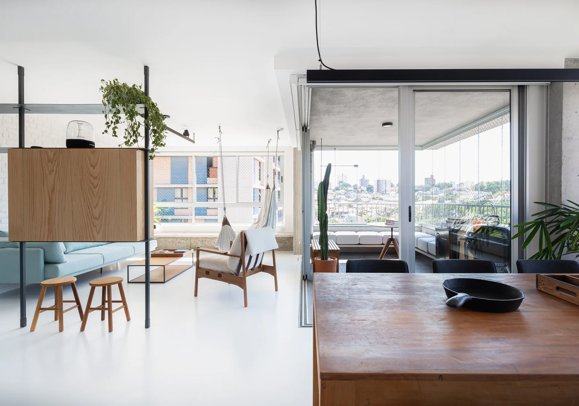 An open-concept living area with white floors features a wooden dining table, a metal-framed room divider, and sliding glass doors leading to a balcony.