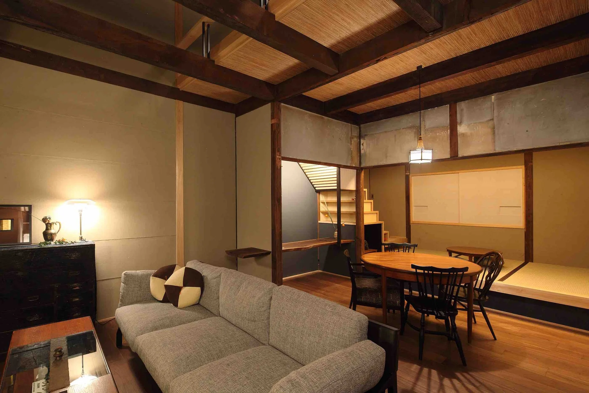A Japanese modern open-plan living room featuring a grey sofa, a wooden dining set, exposed ceiling beams, and a raised tatami platform.