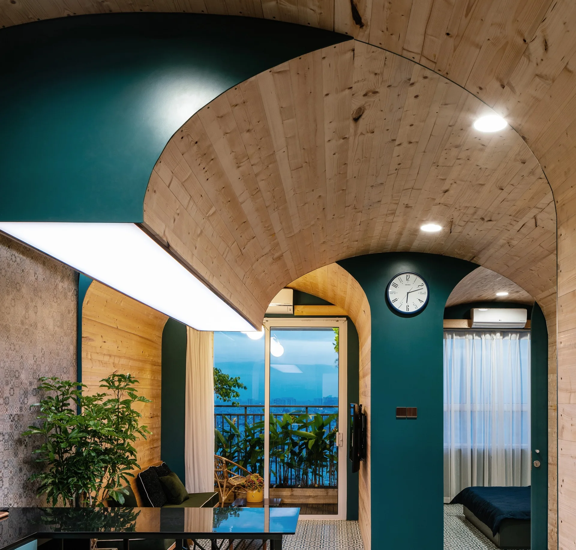 A modern apartment interior featuring light-toned vaulted wood ceilings, deep teal partition walls, patterned tile floors, and a balcony view.