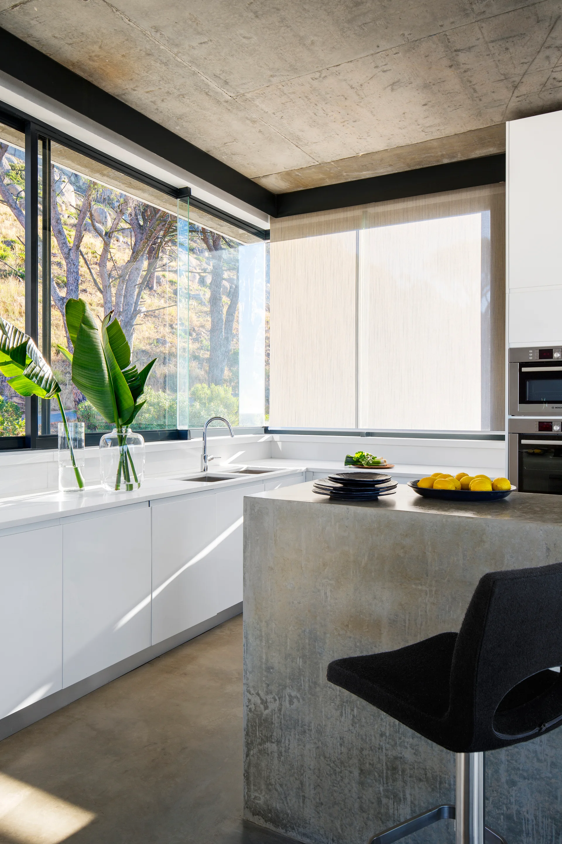 A modern kitchen featuring a raw concrete island, exposed concrete ceiling, white minimalist cabinets, and large corner windows.
