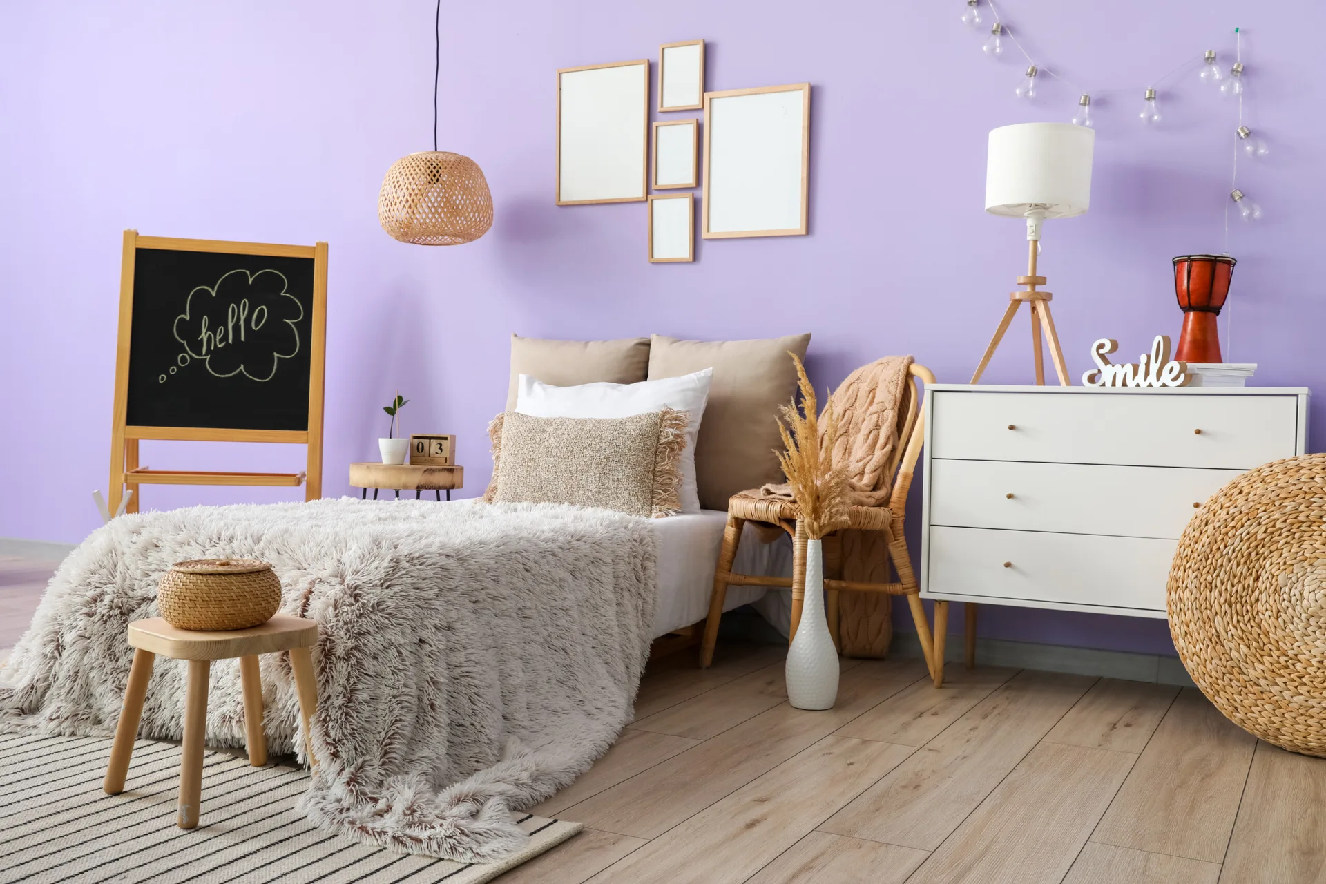 A bedroom with light lavender walls features a bed with neutral linens, a white dresser, wooden frames, and woven rattan accents.